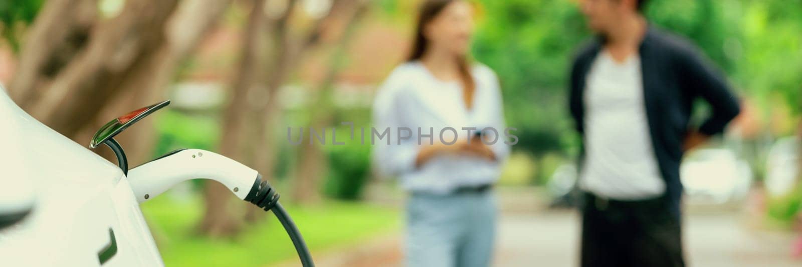 Focused EV car recharging battery on blurred background of lovey couple during autumnal road trip vacation by electric vehicle recharging battery. Eco friendly travel during autumn. Panorama Exalt