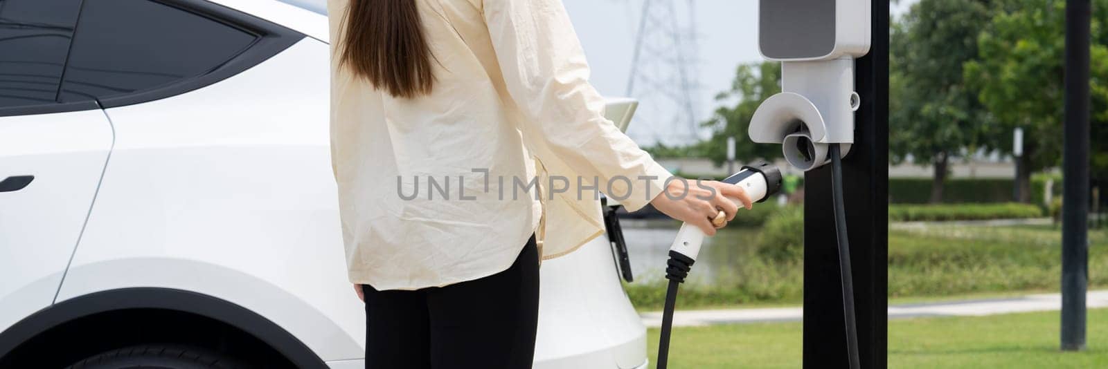 Young woman recharge EV car battery at charging station connected to power grid tower electrical industrial facility as electrical industry for eco friendly vehicle utilization. Panorama Expedient