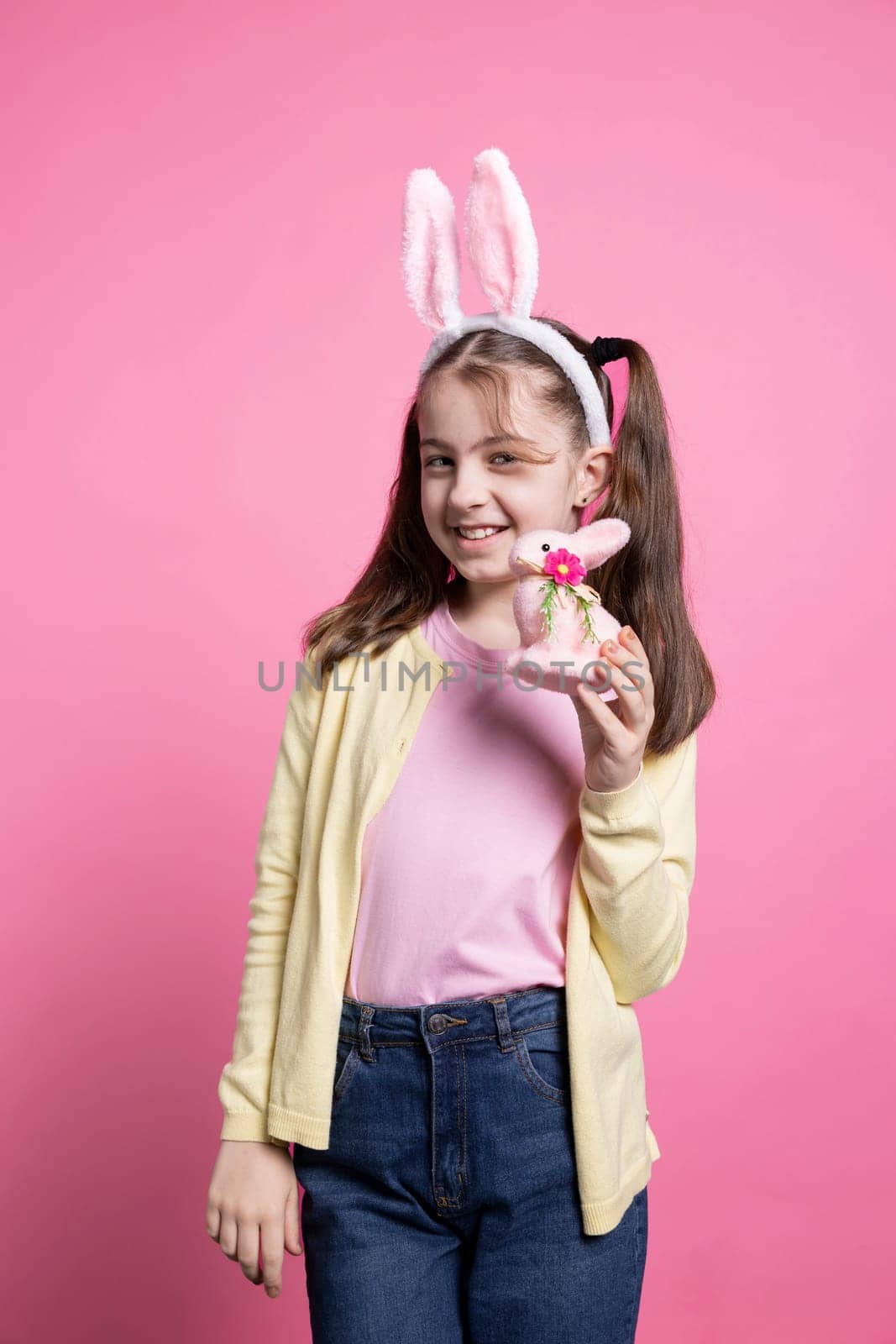 Adorable small kid holding a fluffy pink rabbit toy on camera, presenting her easter colorful toys and ornaments. Young happy child with bunny ears feeling joyful and excited about festivity.