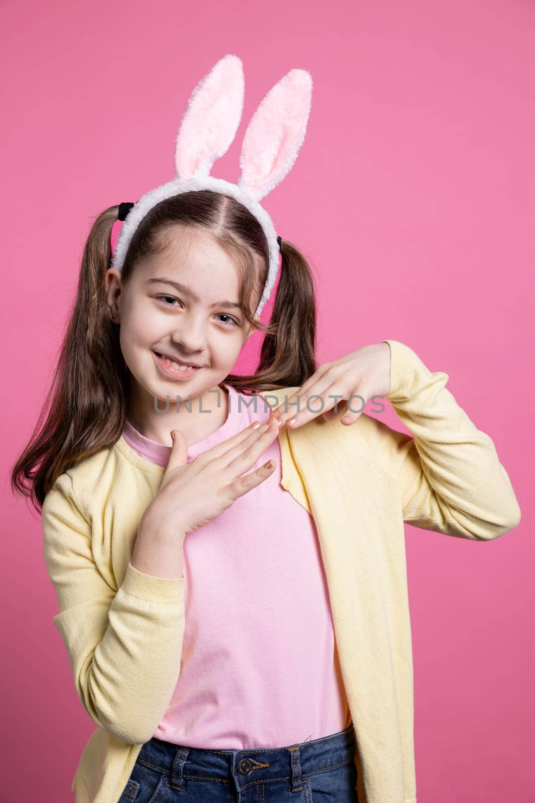Cute young girl dancing and fooling around on camera, wearing bunny ears and pigtails over pink background. Carefree schoolgirl feeling joyful and excited about easter celebration in studio.