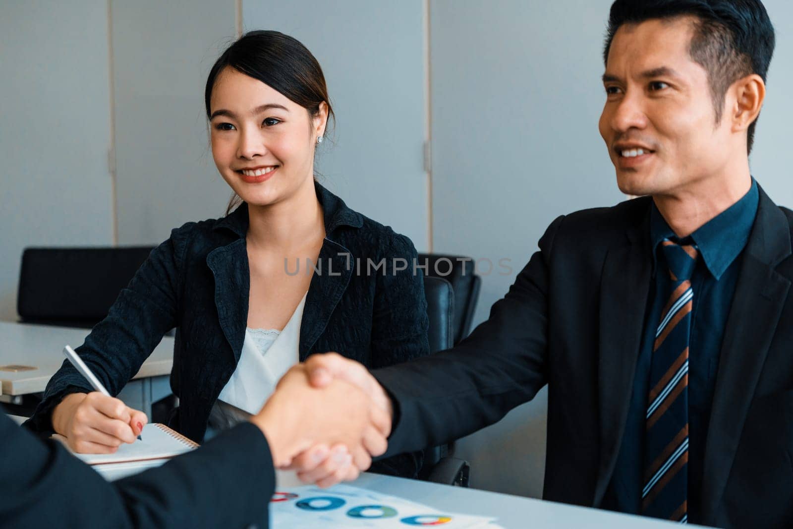 Business people agreement concept. Asian Businessman do handshake with another businessman in the office meeting room. Young Asian secretary lady sits beside him. uds