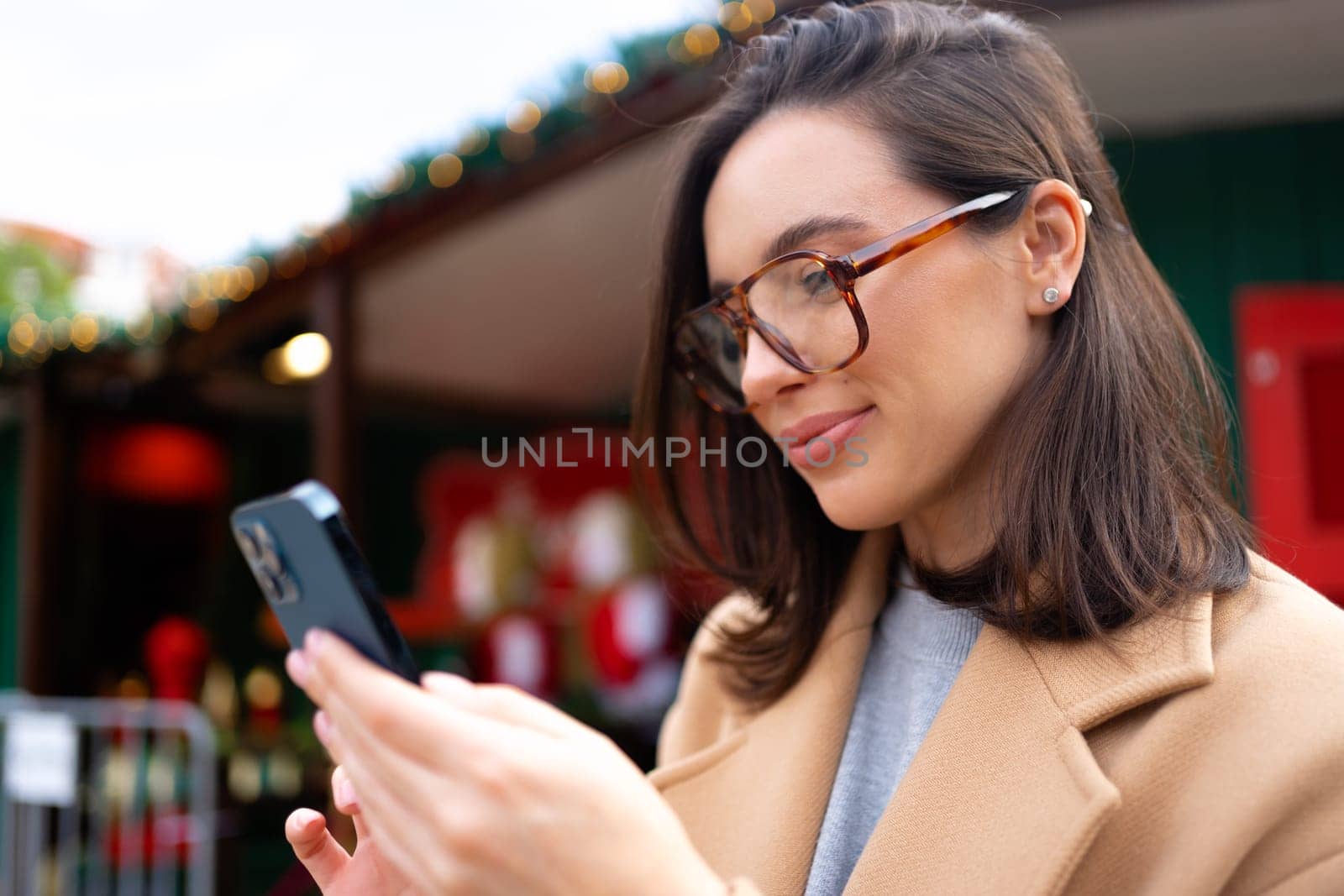 Woman in glasses using mobile phone at day in the city. Happy woman using smartphone dressed stylish trench coat looking device screen