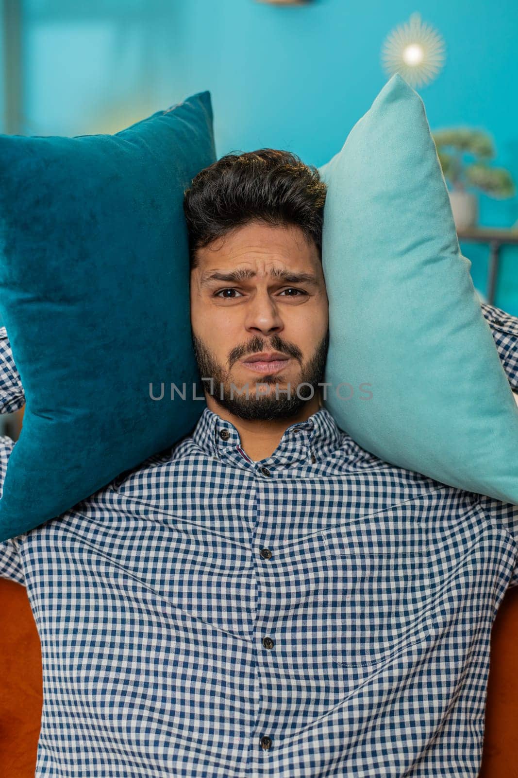 Repair work at neighbors. Irritated Indian Hindu man cover ears with pillows annoyed by noisy loud music neighbors suffer from headache wishes silence. Thin walls at home flat without sound insulation