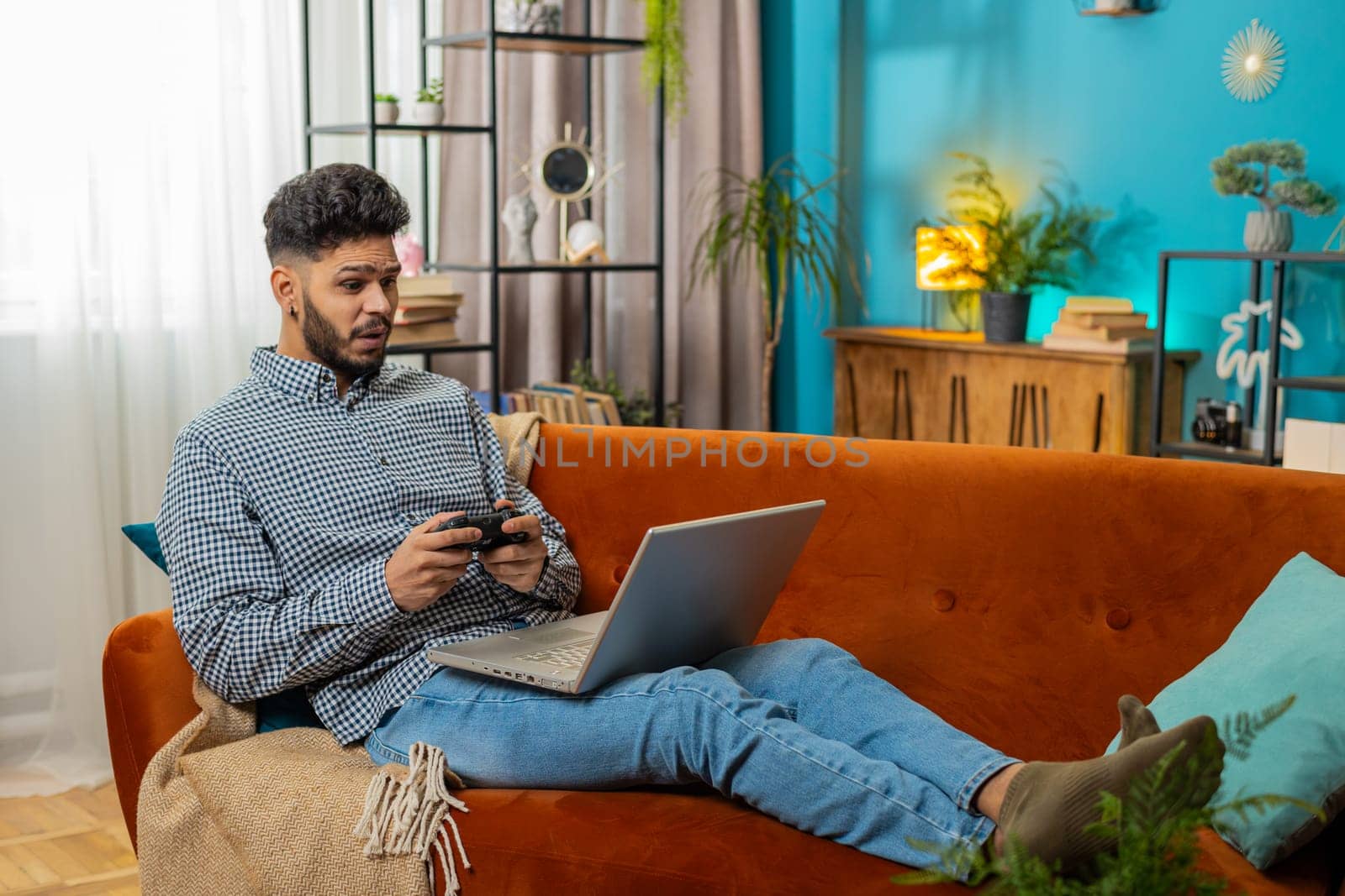 Excited Indian Arabian man playing video game on laptop spending leisure time at home sofa couch. Happy Hispanic freelancer guy playing video games with joystick controller on computer pc. Lifestyle