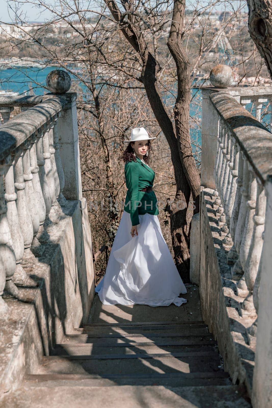 Woman walks around the city, lifestyle. Happy woman in a green jacket, white skirt and hat is sitting on a white fence with balusters overlooking the sea bay and the city. by Matiunina