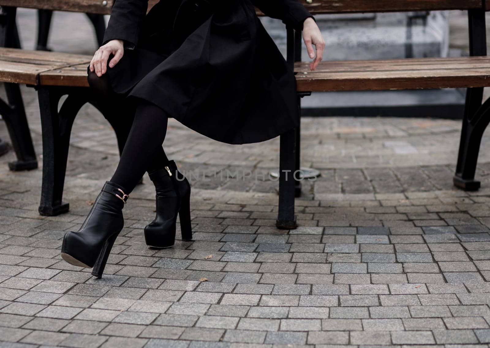 Woman wearing black coat and leather boots and sitting on bench. Trendy casual outfit. Details of everyday look. 