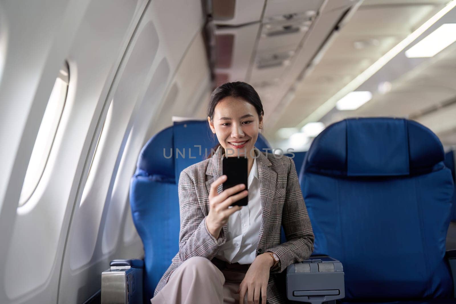 Asian young businesswoman successful or female entrepreneur in formal suit in a plane sit in a business class seat and uses a smartphone during flight. Traveling and Business concept.