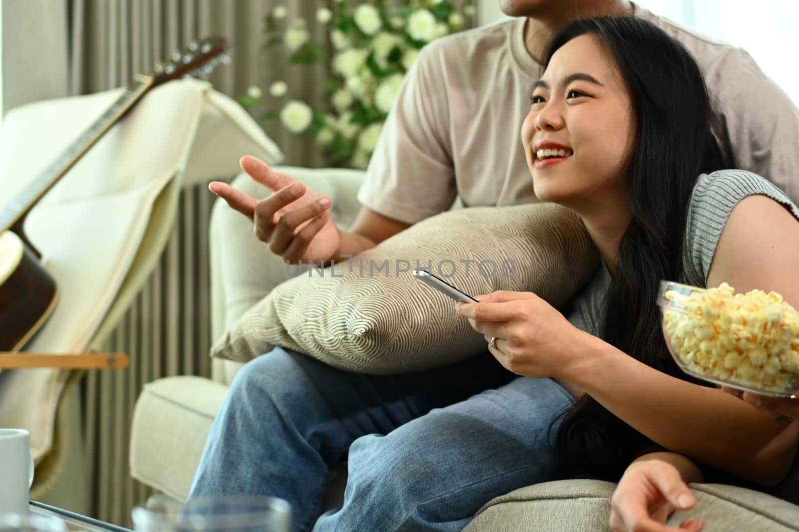 Happy Asian couple eating popcorn and watching movie resting at home on weekend by prathanchorruangsak