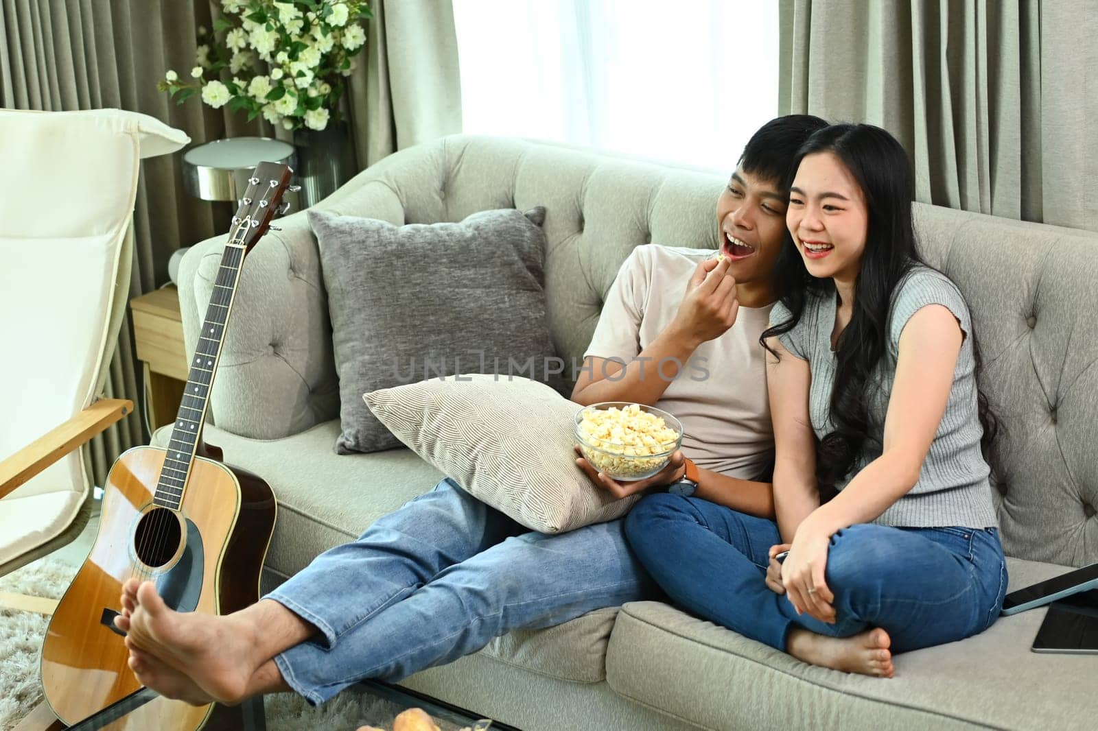 Happy Asian couple eating popcorn and watching movie resting at home on weekend by prathanchorruangsak
