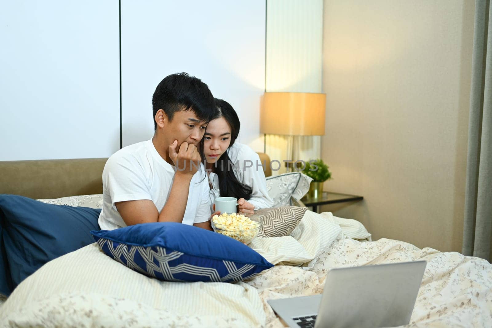 Excited couple watching horror movie on digital tablet during weekend in bedroom.