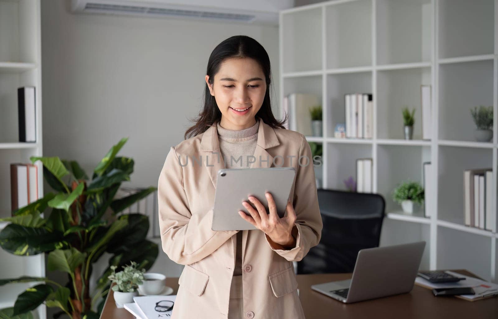 Asian businesswoman holding online business meeting via tablet in modern home office decorated with lush green plants..