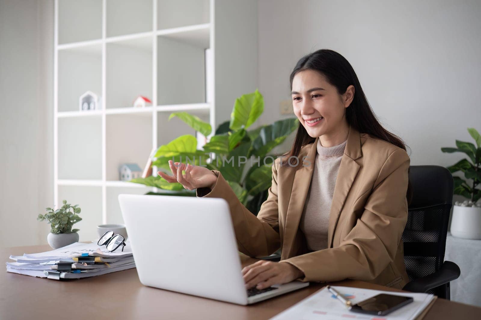 Young Asian business woman sits and works using a laptop in a modern office decorated with shady green plants..