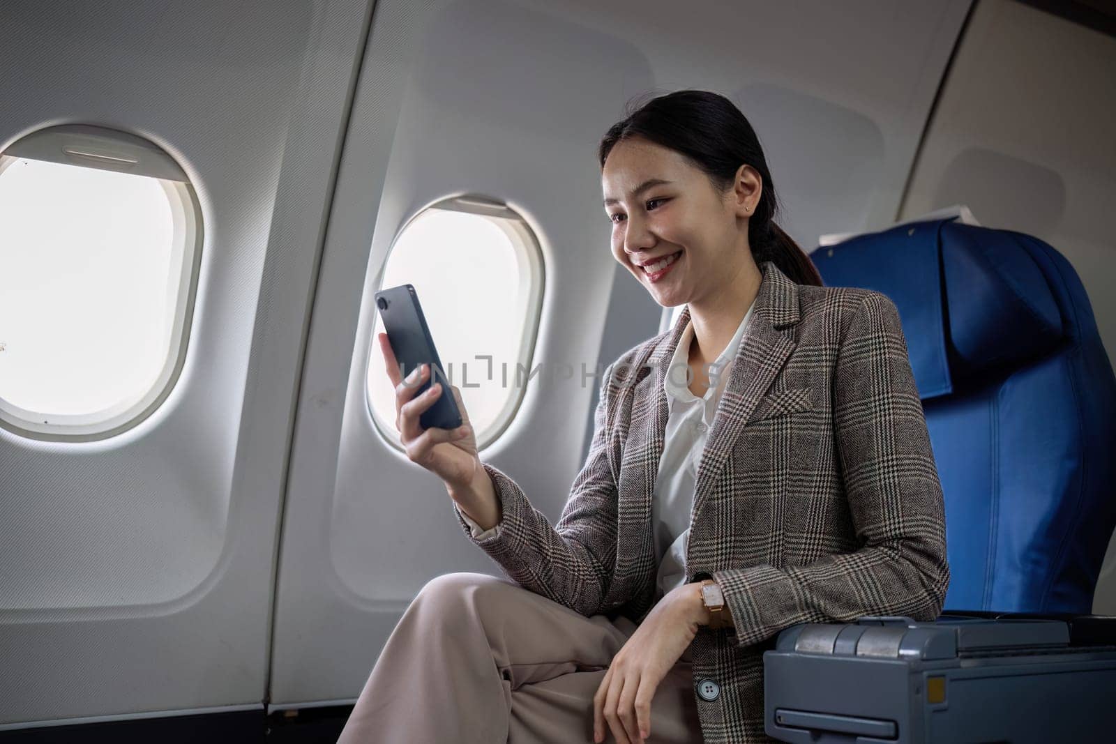 Young Asian woman checks business news on mobile phone, sitting near window in first class on airplane during flight, travel and business concept.