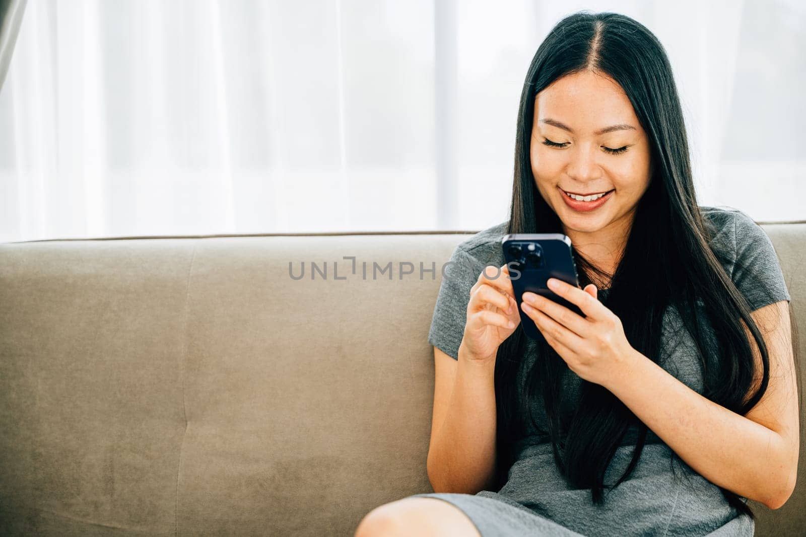 Happy Asian woman sits on a comfortable sofa using her smartphone for texting chatting and online shopping. Enjoying technology for connection and leisure at home.