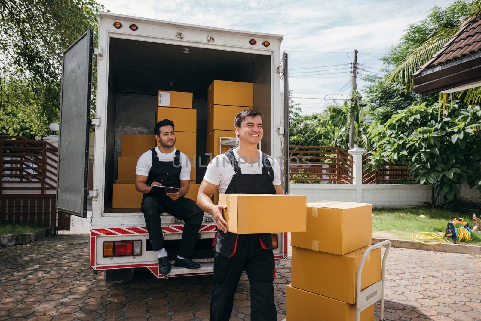 Moving service workers unload boxes from a van showing teamwork and cooperation. Delivery men in uniform relocating items. Smiling employees carrying orders. relocation teamwork Moving Day by Sorapop