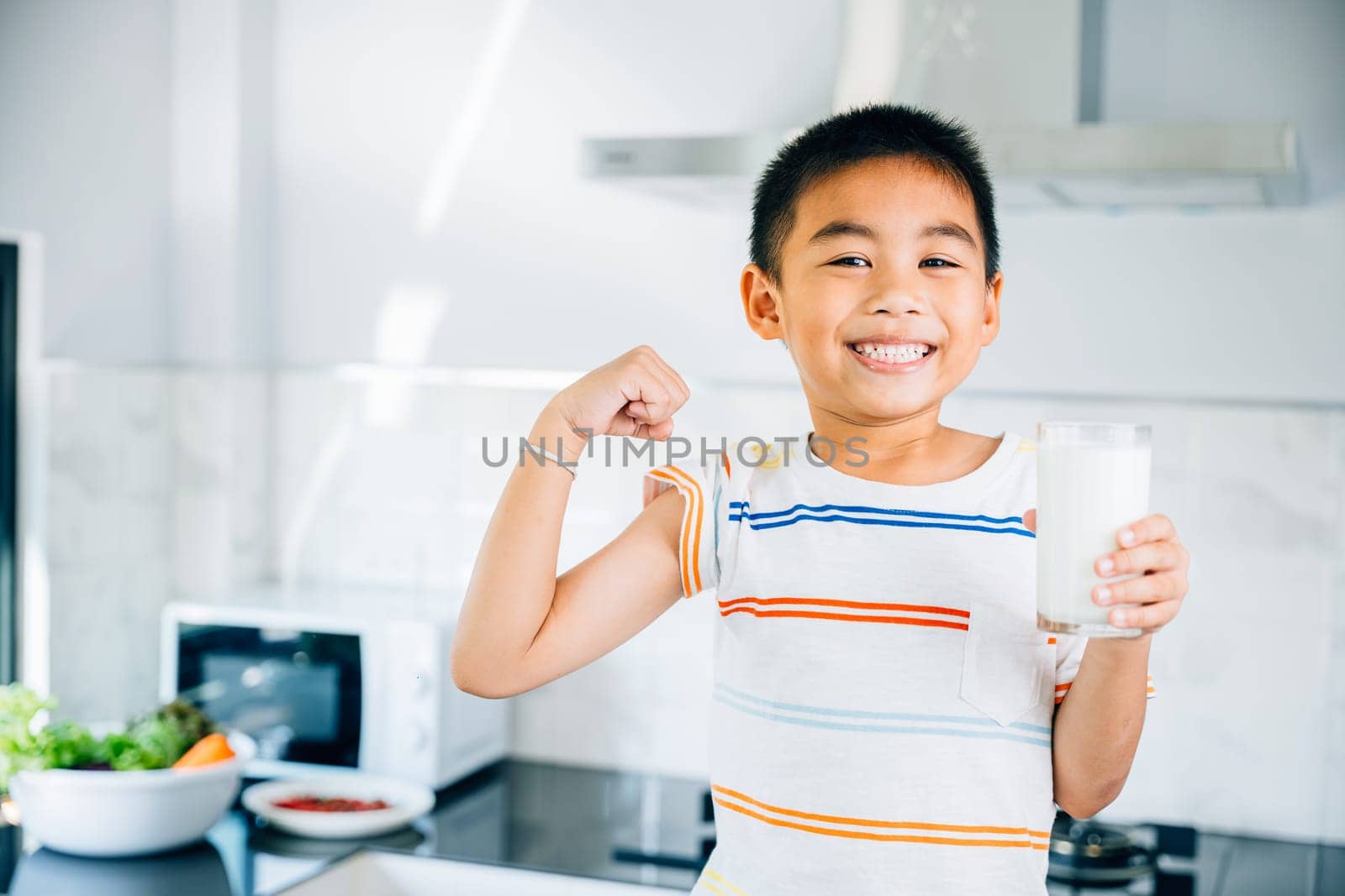 Cheerful Asian little boy holds milk in kitchen, smiling brightly. Portrait of cute son enjoying drink. Joyful child sips calcium-rich liquid, radiating happiness at home give me.