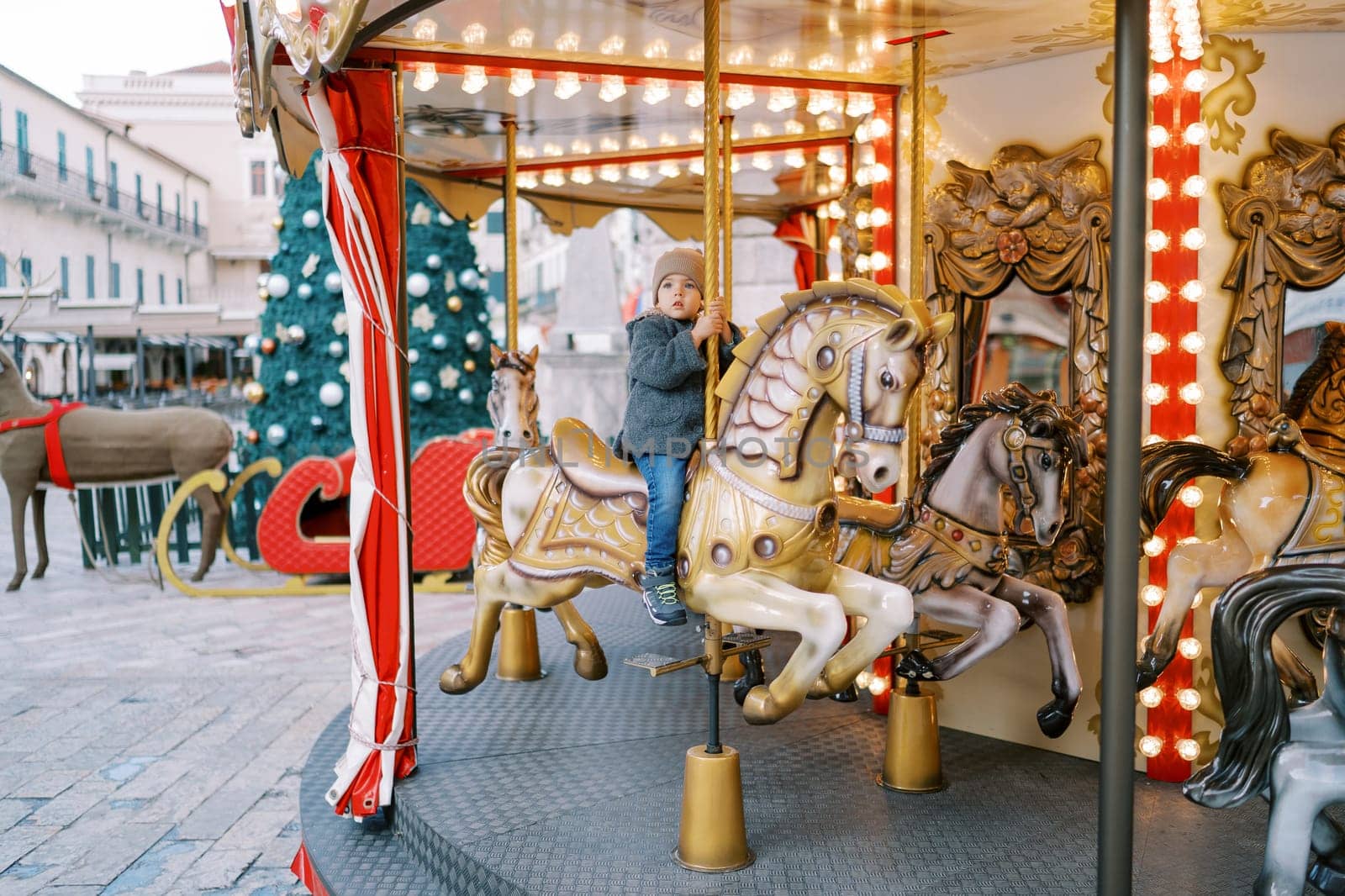 Little girl rides a toy horse on a carousel at a fair near a decorated Christmas tree near an old house. High quality photo