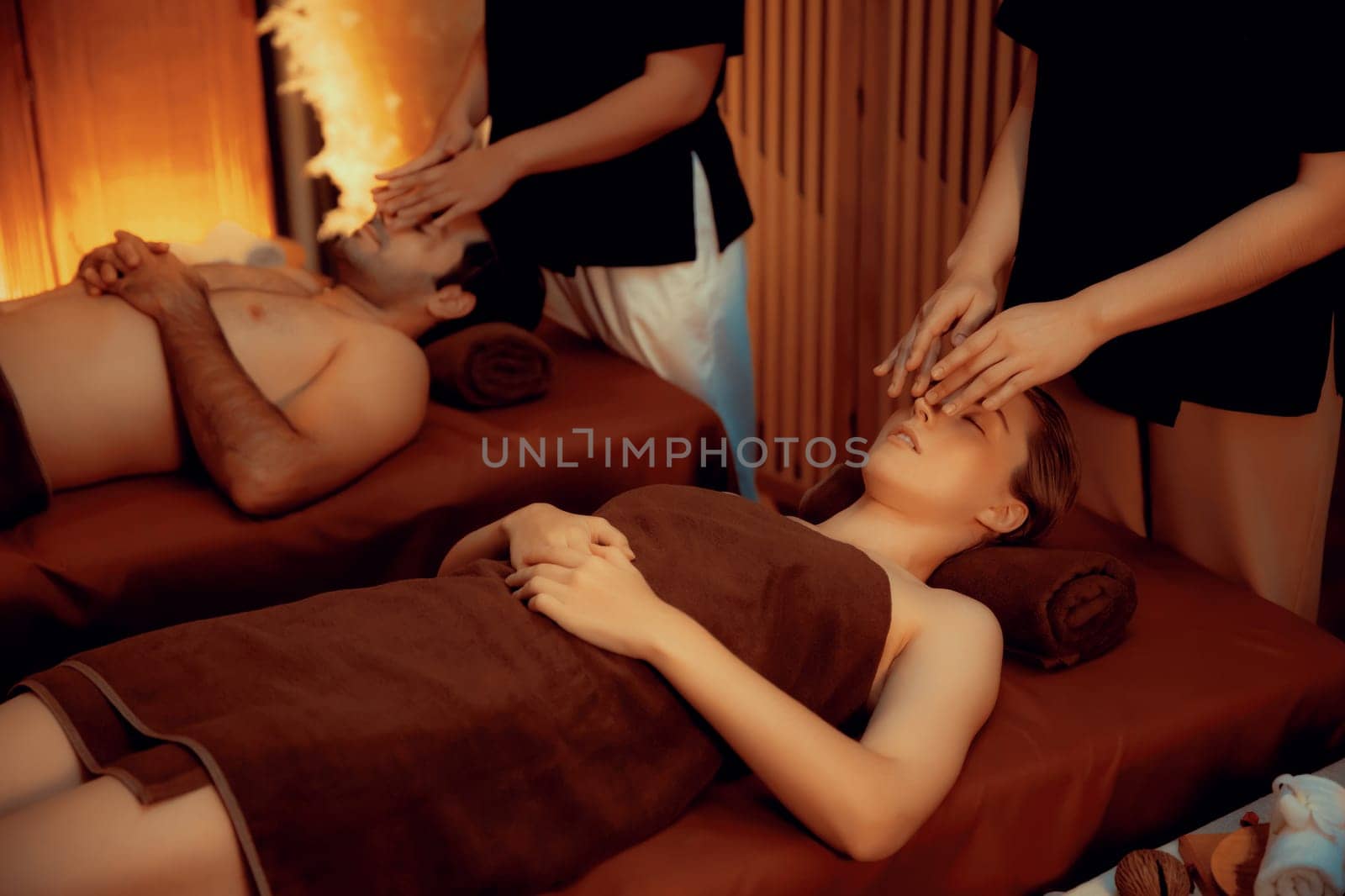 Couple customer enjoying relaxing anti-stress head massage and pampering facial beauty skin recreation leisure in warm candle lighting ambient salon spa in luxury resort or hotel. Quiescent