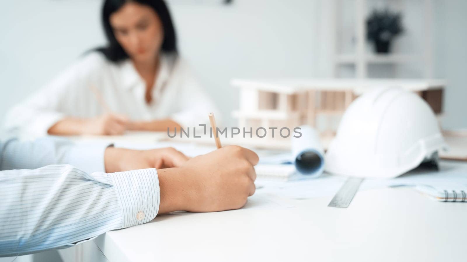 Professional architect engineer drawing blueprint and working together with young beautiful caucasian coworker on meeting table with safety helmet, house model, blueprint scatter around. Immaculate.