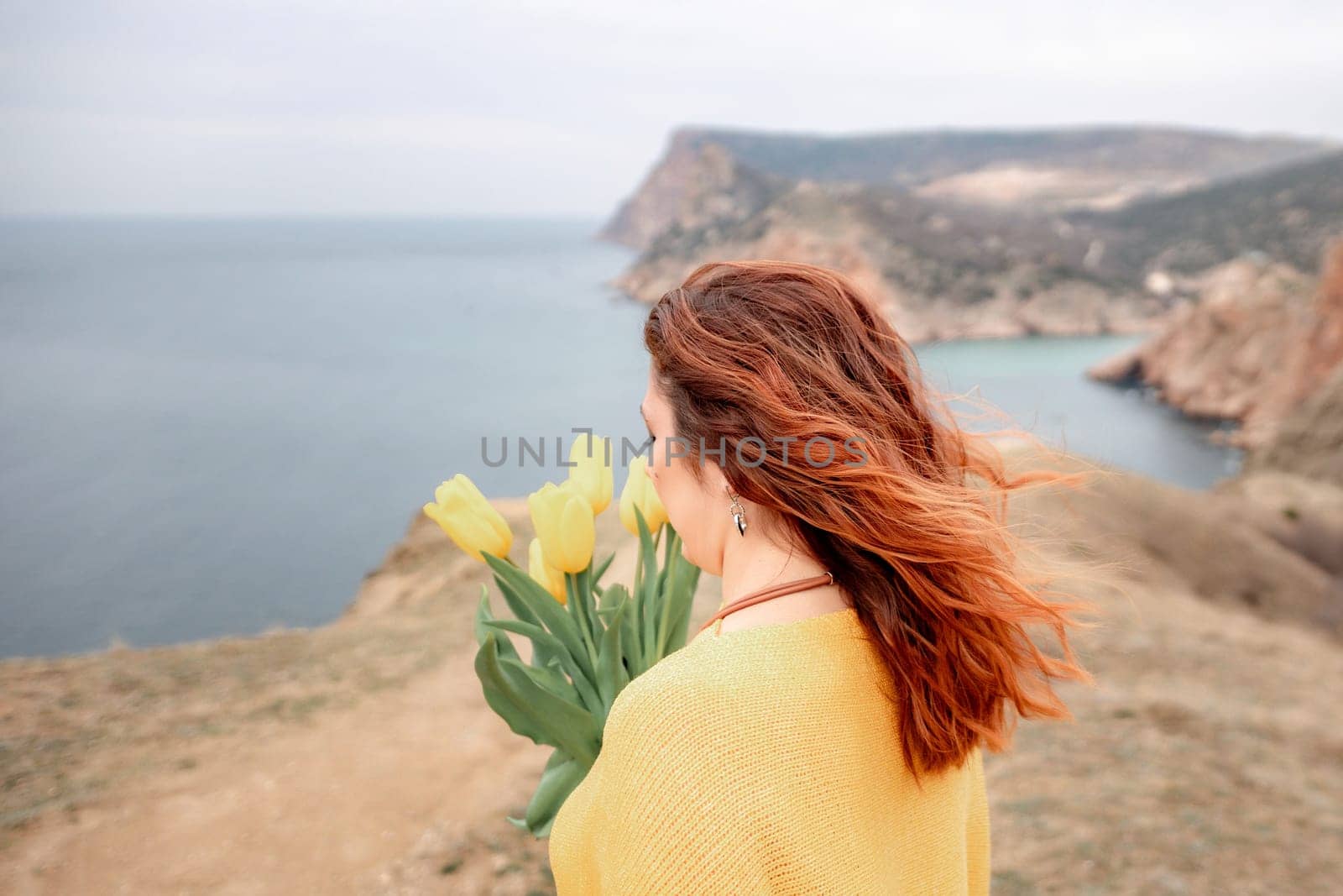 Rear view of a woman with long hair against a background of mountains and sea. Holding a bouquet of yellow tulips in her hands, wearing a yellow sweater.