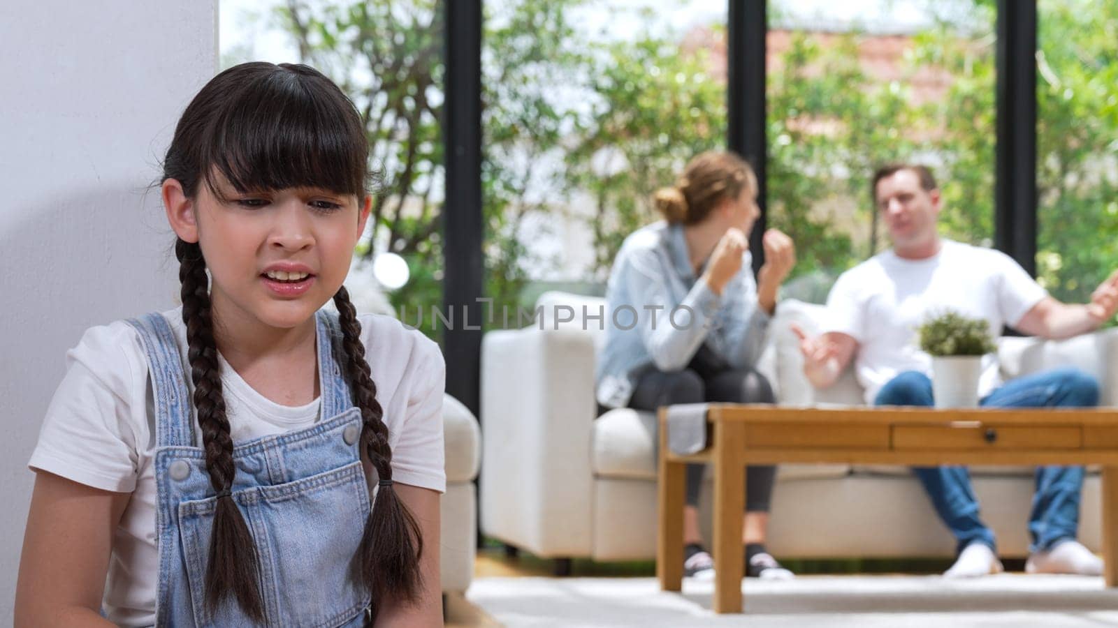 Stressed and unhappy young girl huddle in corner, cover her ears blocking sound of her parent arguing in background. Domestic violence at home and traumatic childhood develop to depression. Synchronos