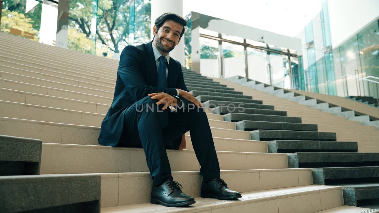 Smiling skilled businessman looking at camera while sitting on stairs. Young professional project manager smile at camera while holding mobile phone at outdoor with blurred background. Exultant.