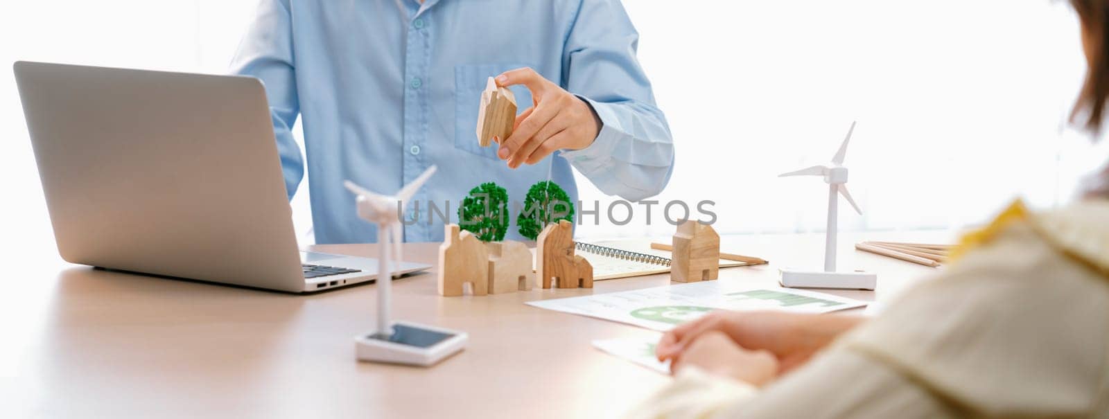 Two business people talking about eco city at green business meeting on meeting table with windmill represented renewable energy, wooden house block and tree model represent green city. Delineation.