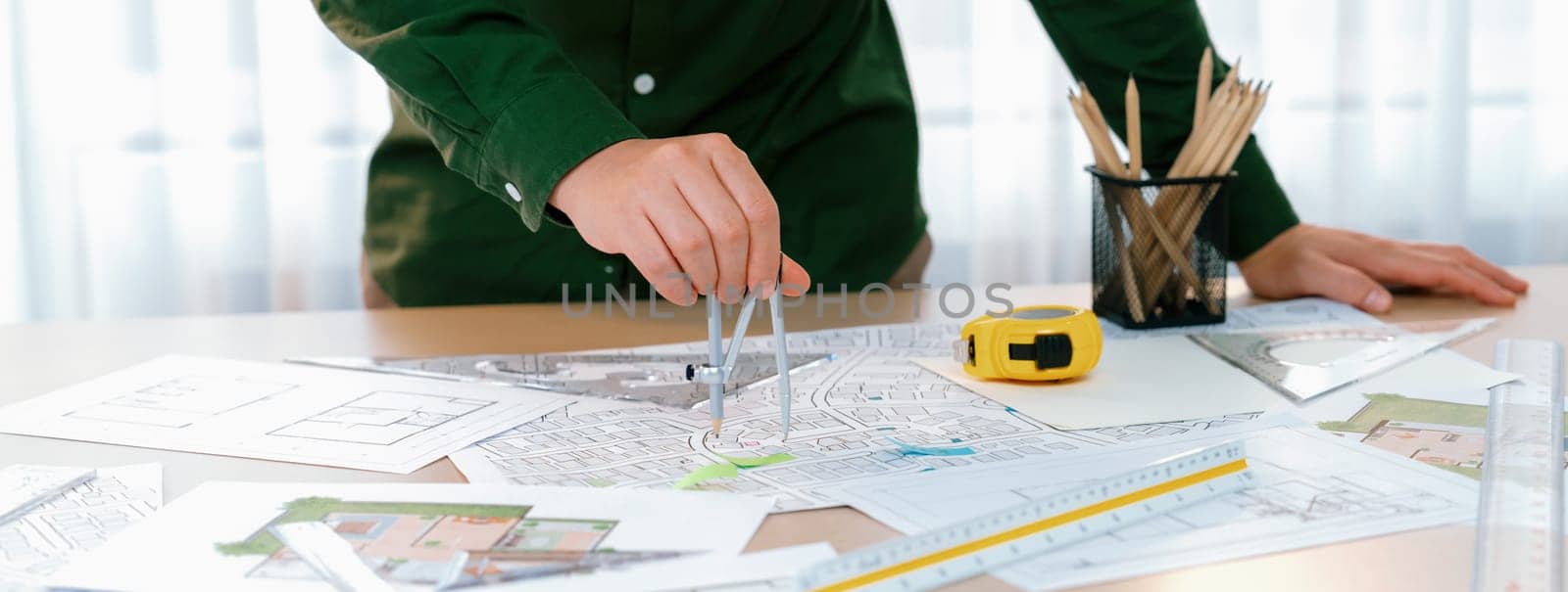 A portrait of architect using divider to measure blueprint. Architect designing house construction on a table at studio, with architectural equipment scattered around. Focus on hand. Delineation