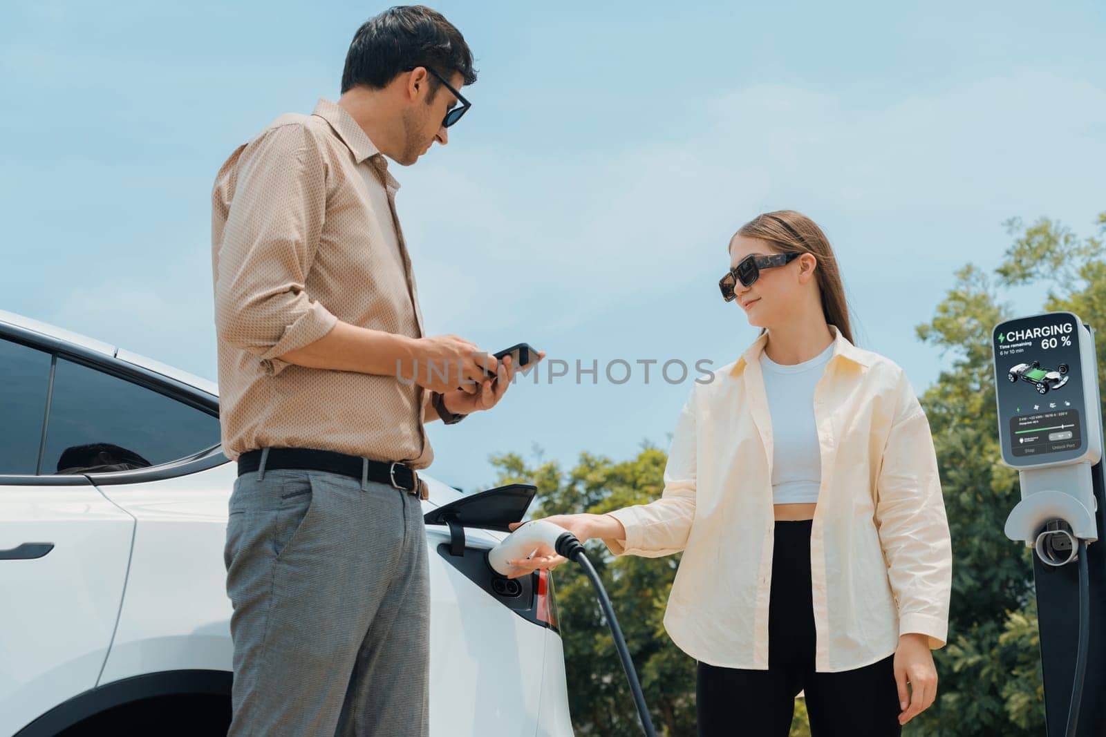 Young couple use smartphone to pay for electricity at public EV car charging station green city park. Modern environmental and sustainable urban lifestyle with EV vehicle. Expedient