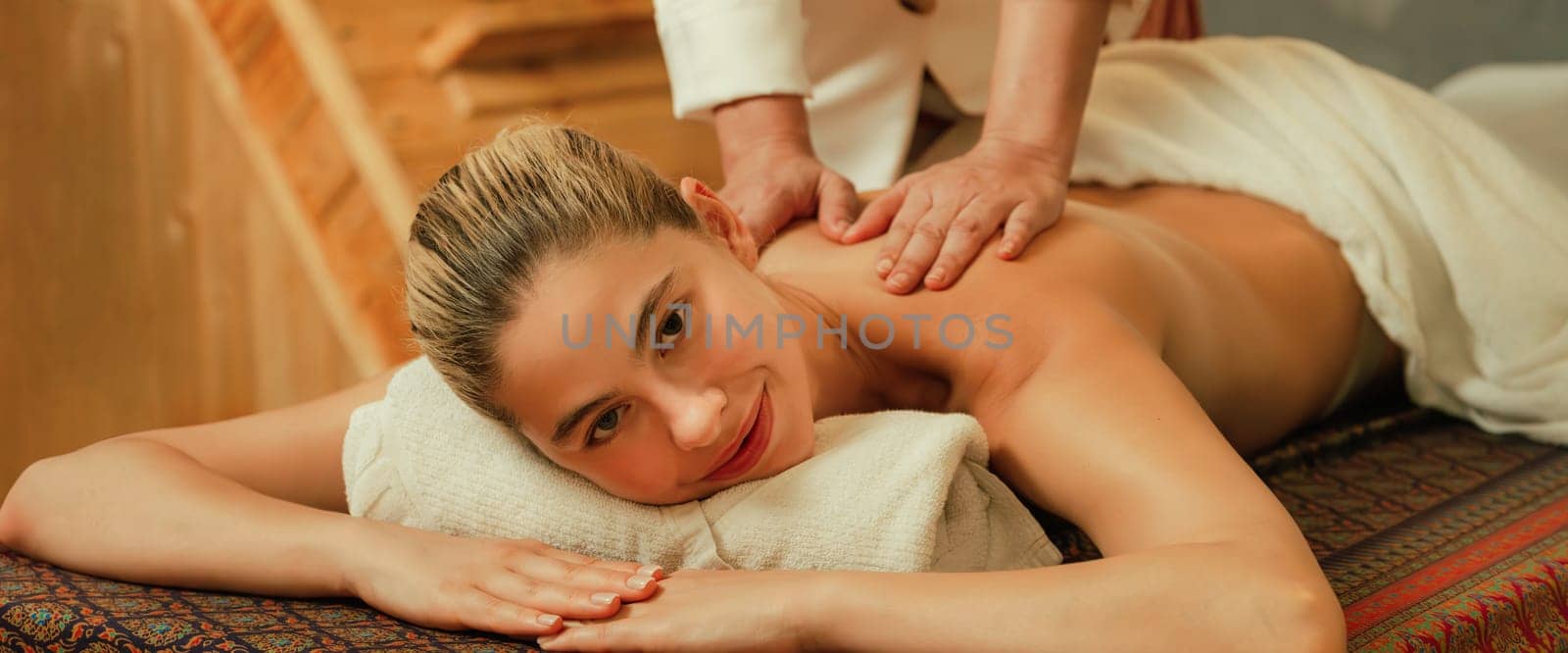 A portrait of a pretty girl having body massage while looking at camera with happiness in front of wooden sauna cabinet. Attractive blonde female feel relaxed from Thai massage. Tranquility.