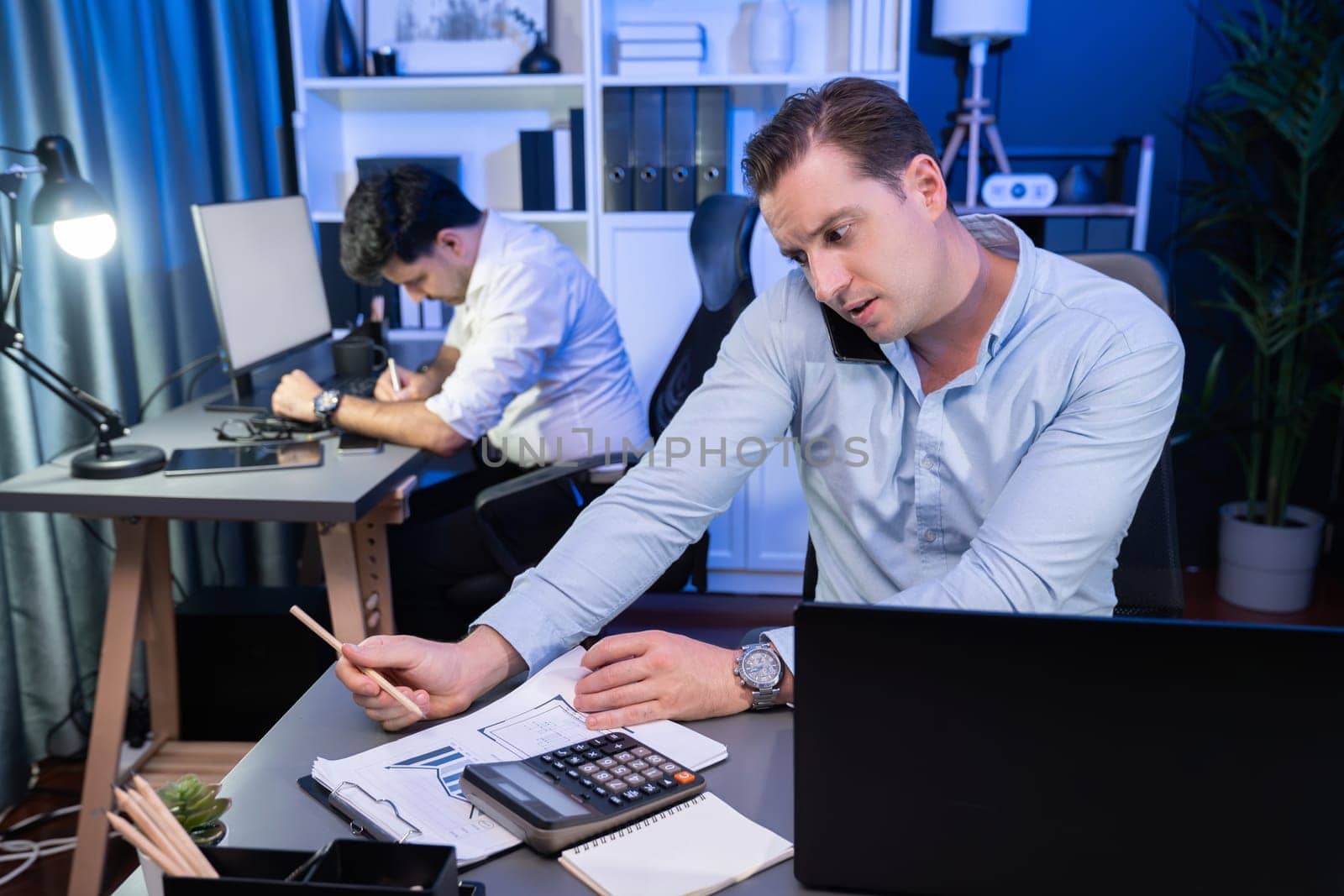 Serious businessman calling on phone with customer to create new project at night time, coworker writing document work sheet on desk. Concept of working on neon blue light modern office. Sellable.