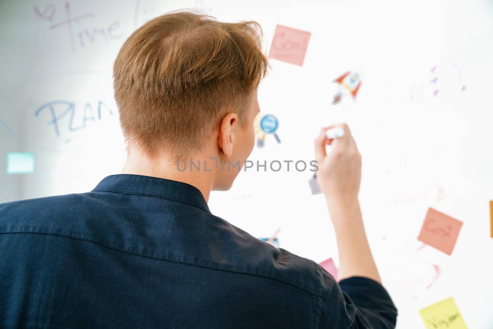 Professional caucasian businessman writing and sharing marketing idea by using mind map and sticky notes on glass board at meeting room. Creative business and planing concept. Back view. Immaculate.