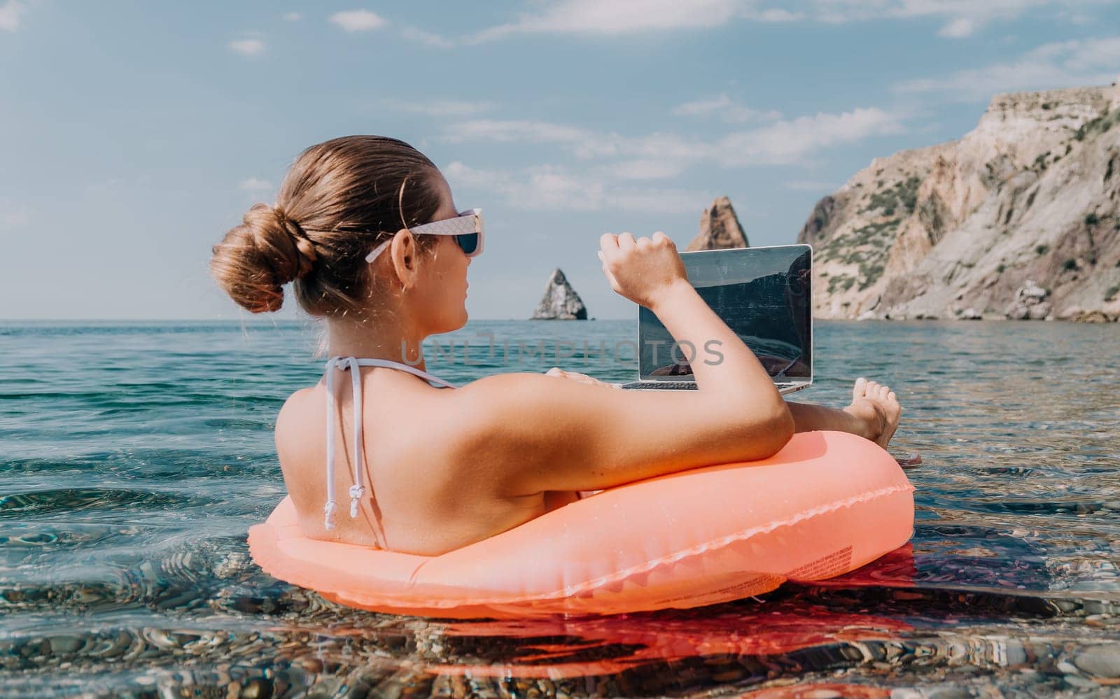 Woman freelancer works on laptop swimming in sea on pink inflatable ring. Pretty lady typing on computer while floating in the sea on inflatable donut at sunset. Freelance, remote work on vacation