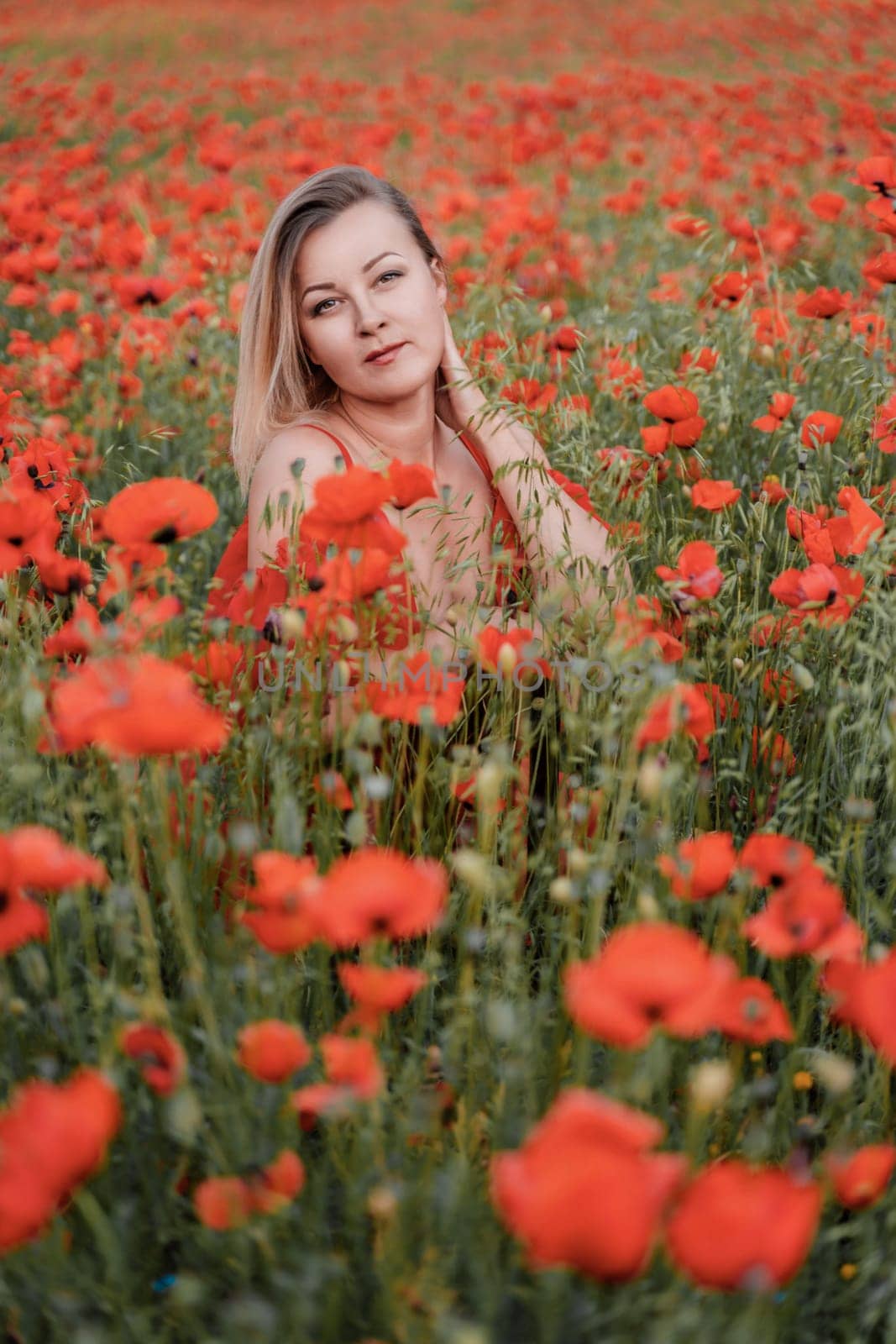 Happy woman in a red dress in a beautiful large poppy field. Blond sits in a red dress, posing on a large field of red poppies.