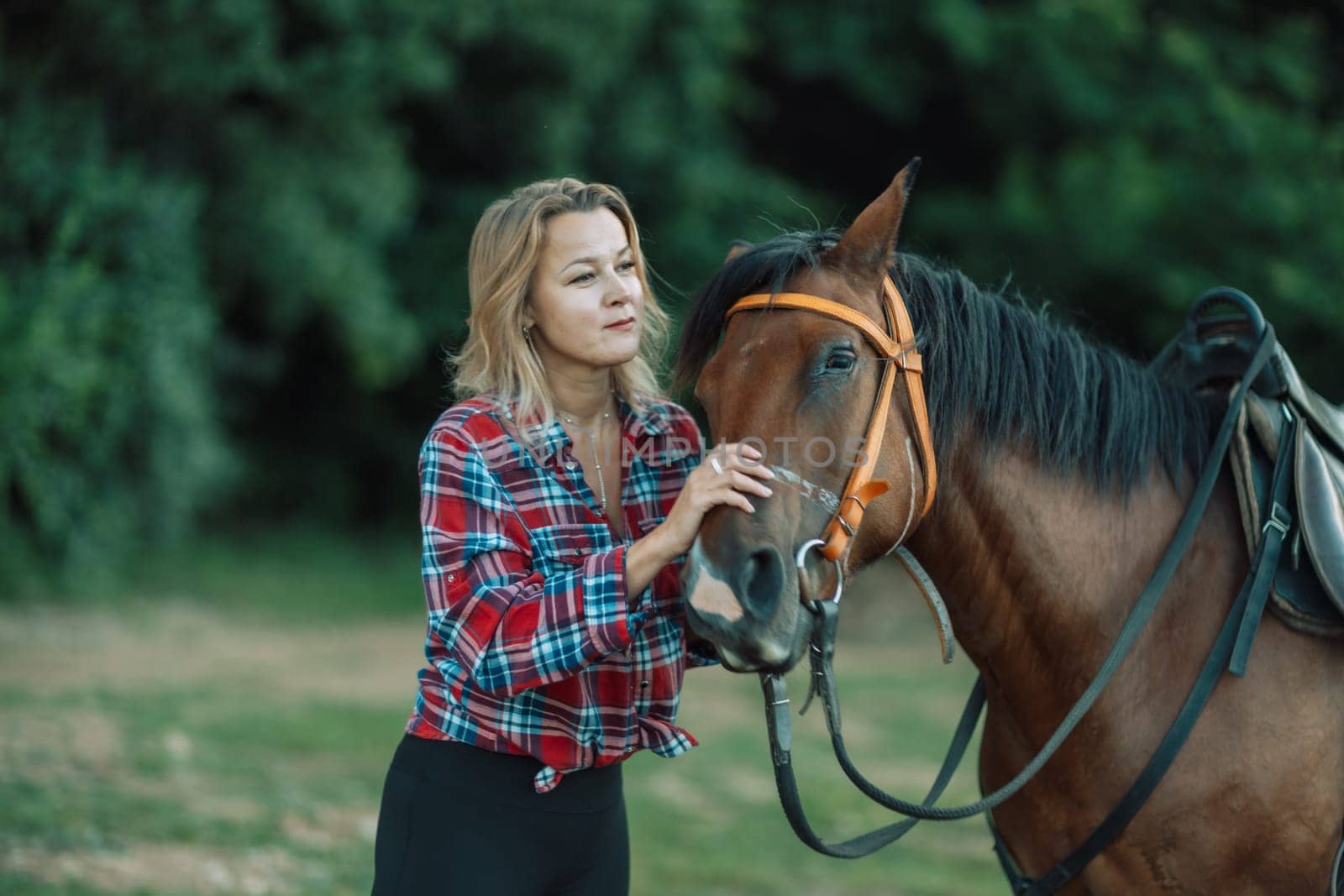 Happy blonde with horse in forest. Woman and a horse walking through the field during the day. Dressed in a plaid shirt and black leggings