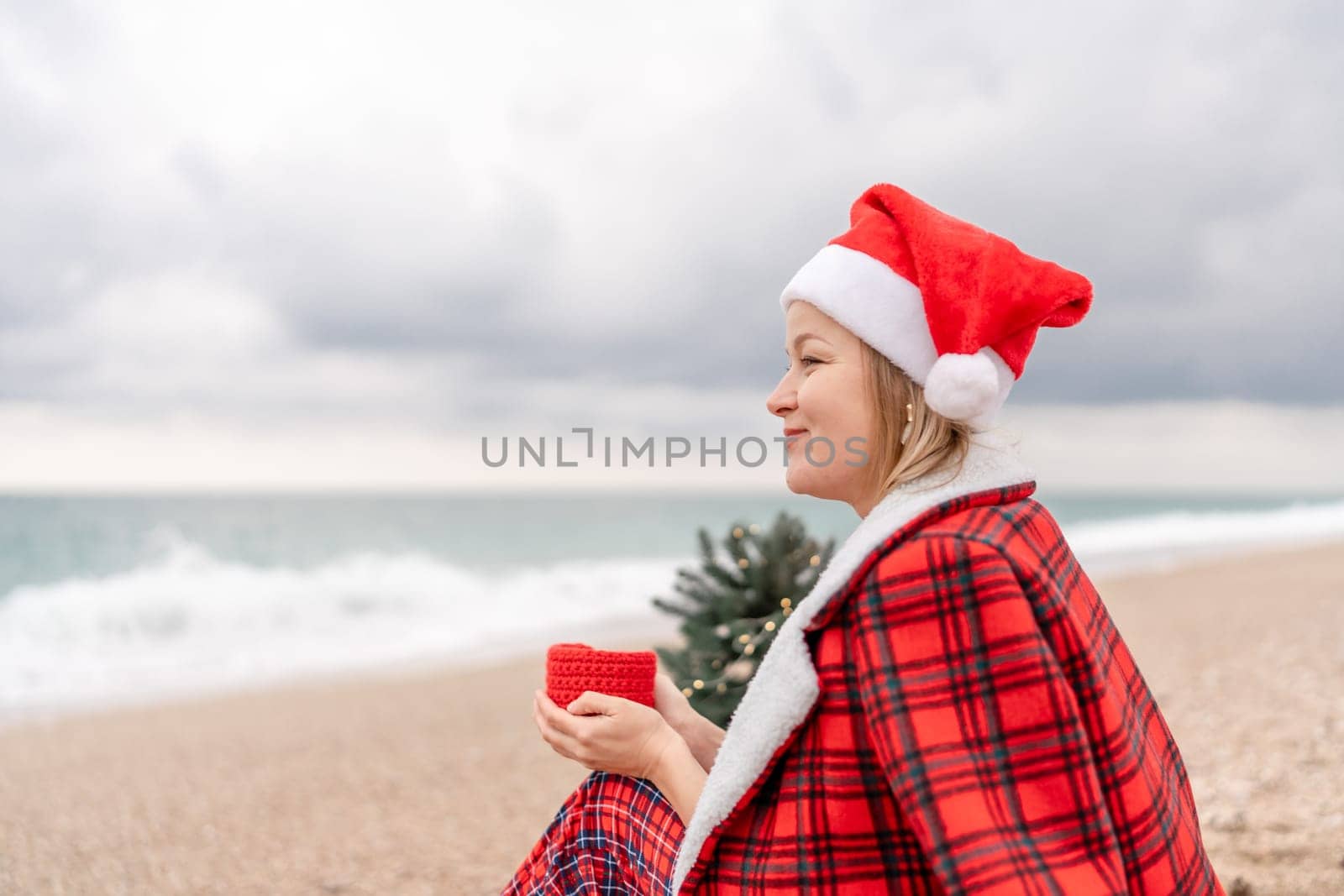 Sea Lady in Santa hat plaid shirt with a red mug in her hands enjoys beach with Christmas tree. Coastal area. Christmas, New Year holidays concep.