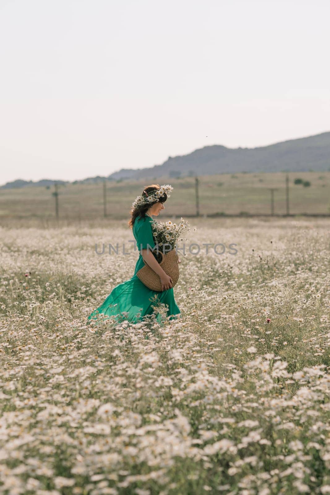 Happy woman in a field of daisies with a wreath of wildflowers on her head. woman in a green dress in a field of white flowers. Charming woman with a bouquet of daisies, tender summer photo.