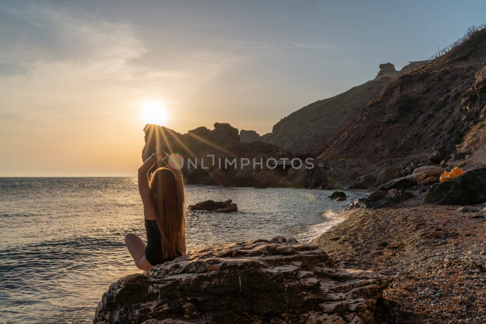Woman tourist enjoying sunset over the sea mountain landscape. Sits outdoors on a rock above the sea. She is wearing jeans and a blue hoodie. Healthy lifestyle, harmony and meditation.