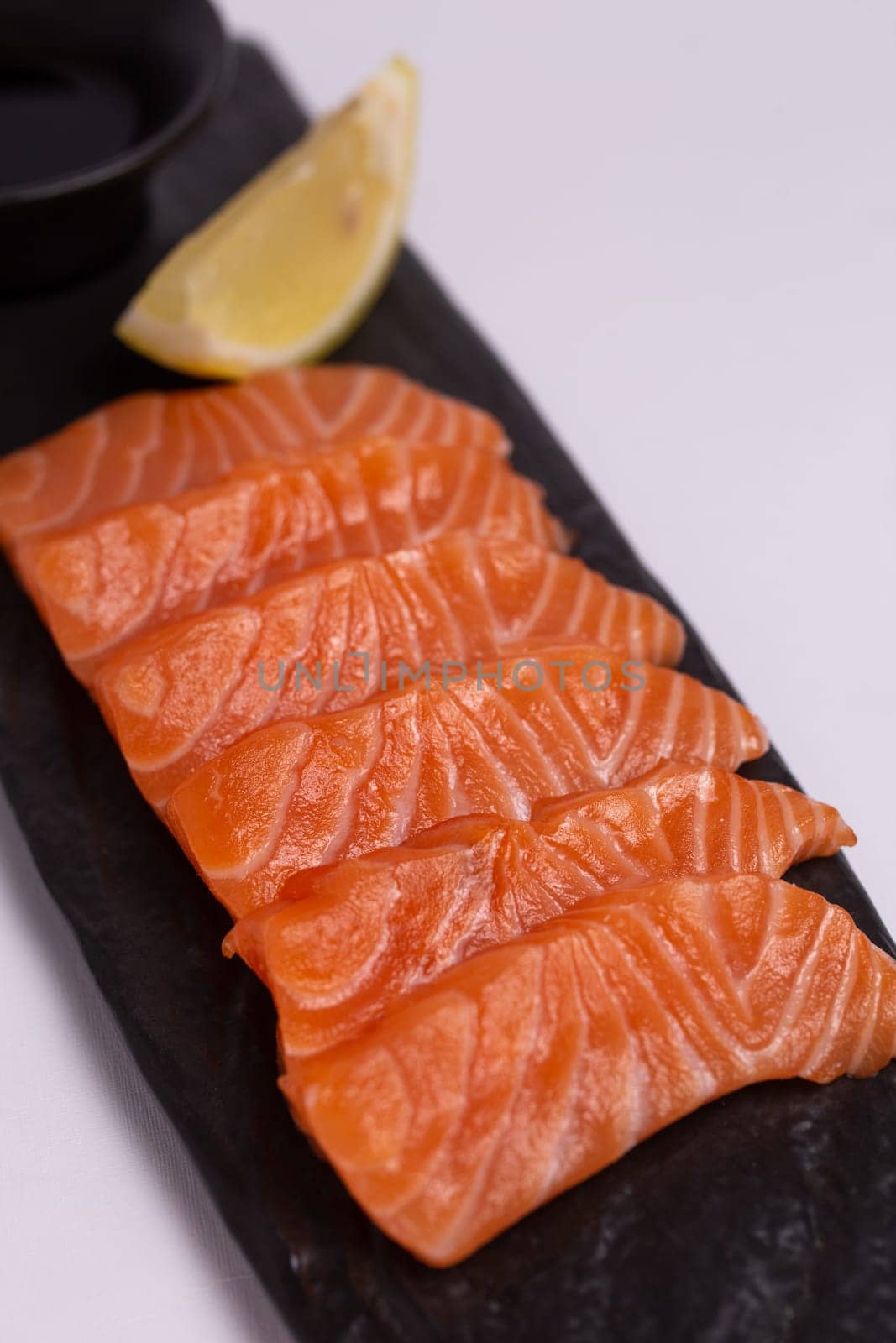 A close-up of a fresh salmon fillet garnished with lemon wedges and soy sauce on a black stone plate. This appetizing seafood dish is isolated on a white background, perfect for a healthy meal.