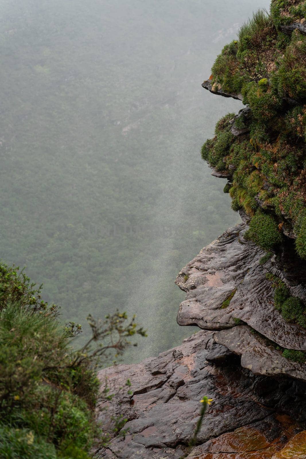 A waterfall's top part is blown by winds, forming a misty curtain above a verdant valley.