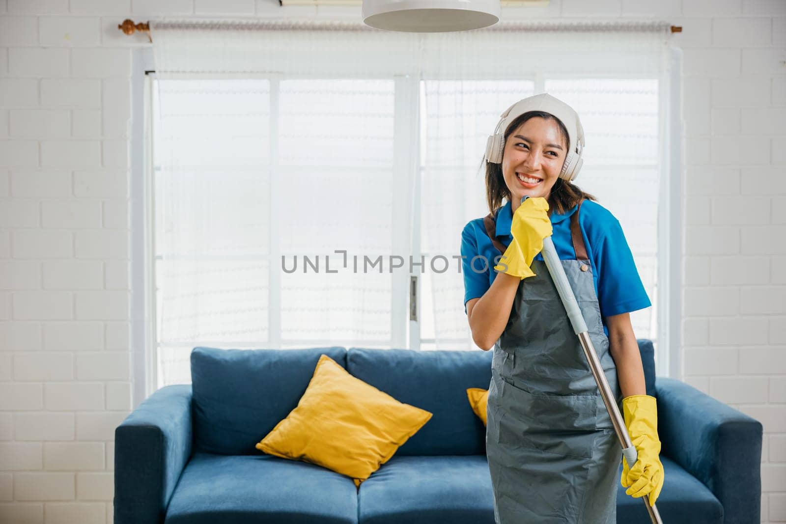 Happy Asian housewife sings her favorite song while cleaning using mop as microphone for fun during domestic work. Dancing and cleaning in living room cheerful maid enjoying her routine. by Sorapop