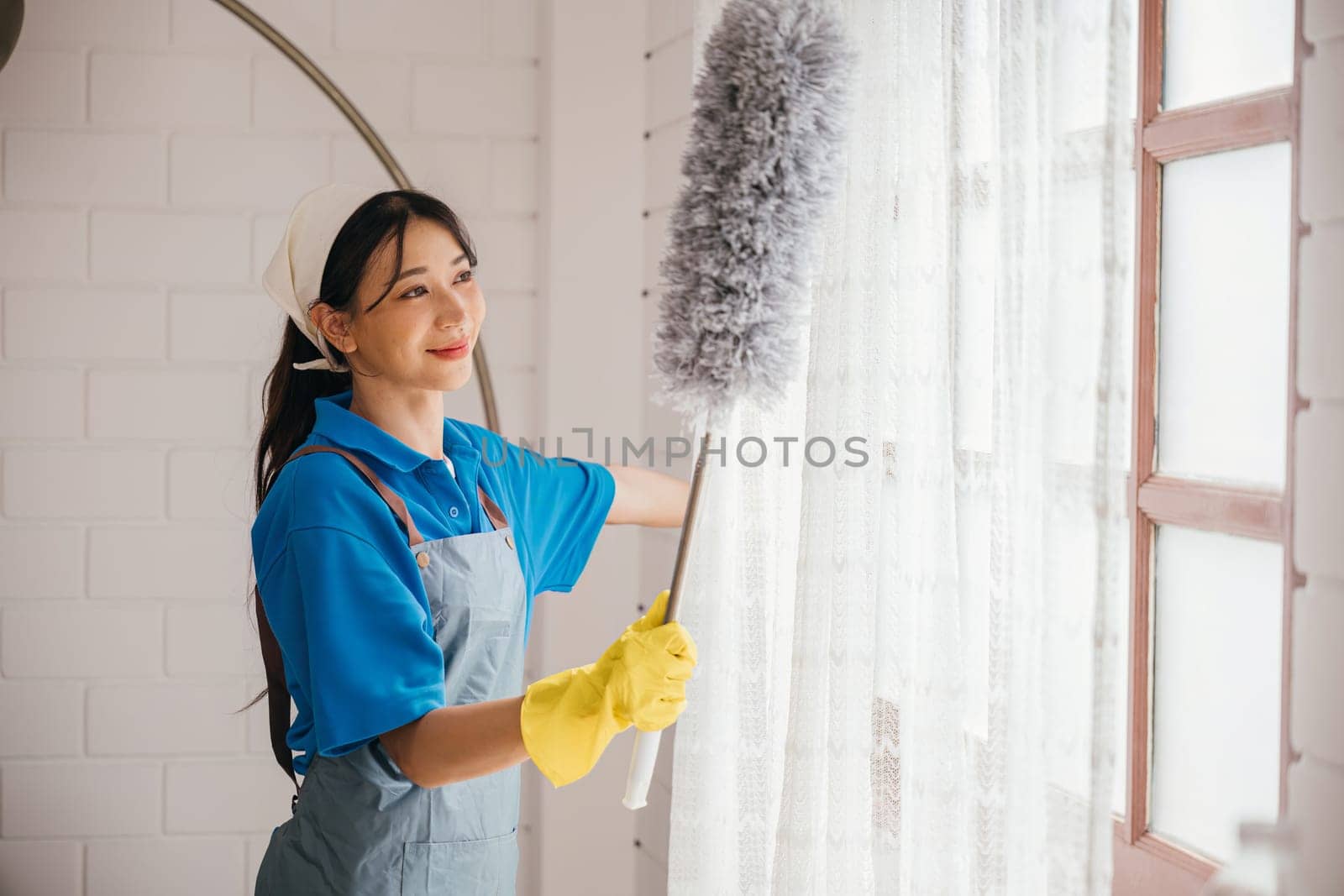Asian woman's portrait while dusting window blinds. She stands holding duster smiling happily. Occupation involves house hygiene routine cleaning and care. Cleaning service for modern clean household.