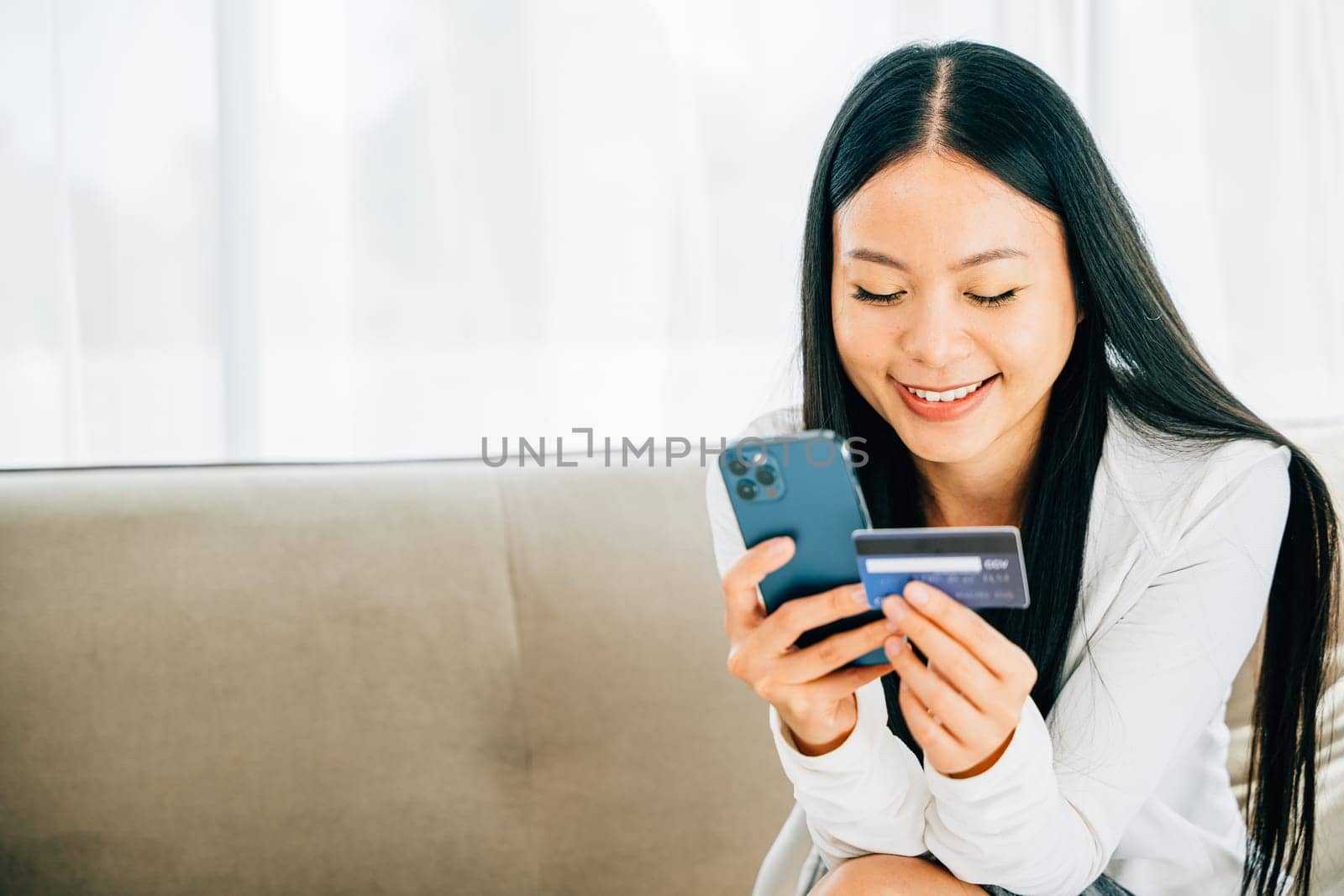 Smiling woman holds smartphone and credit card shops online on sofa by Sorapop