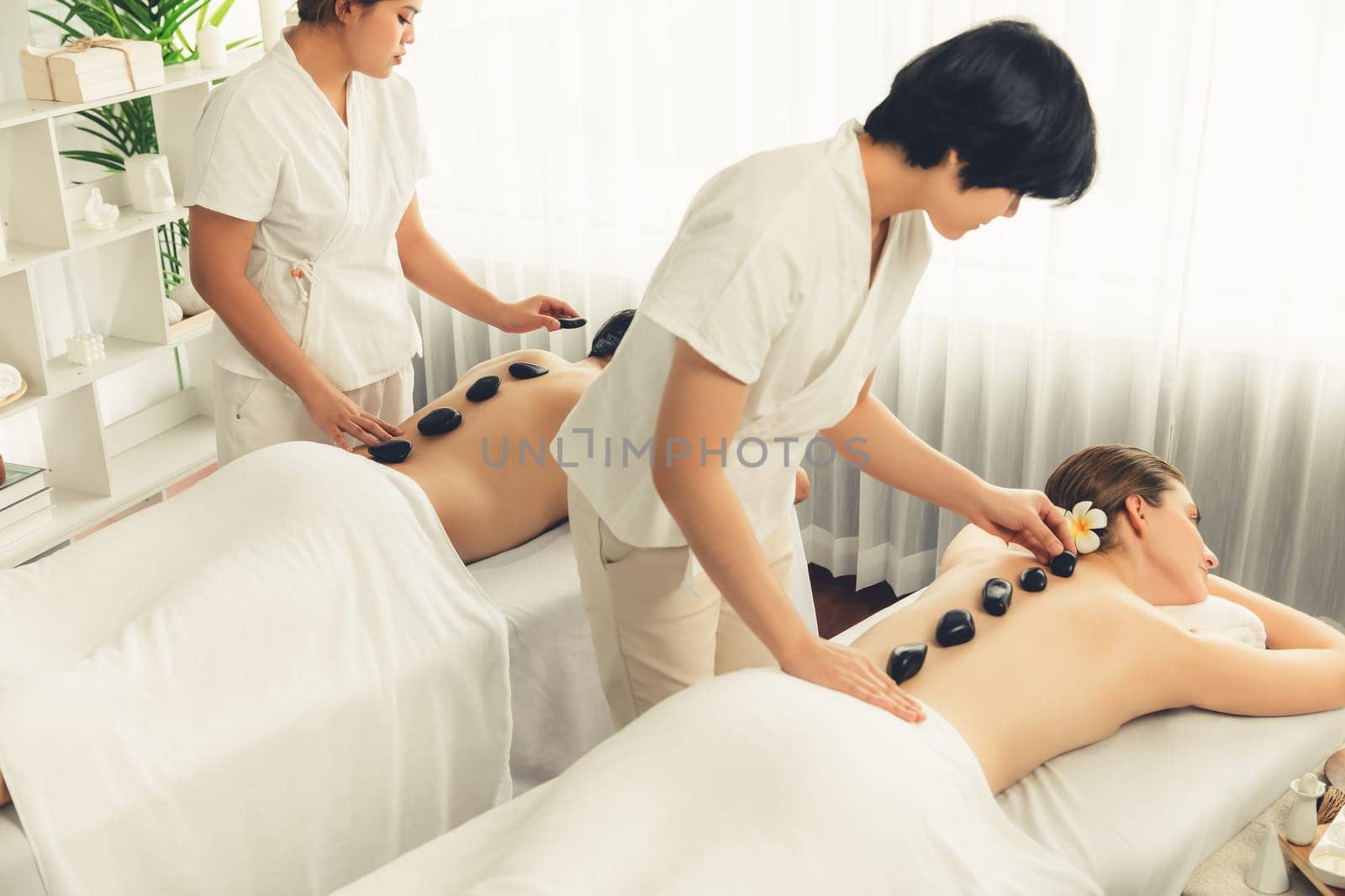 Hot stone massage at spa salon in luxury resort with day light serenity ambient, blissful couple customer enjoying spa basalt stone massage glide over body with soothing warmth. Quiescent