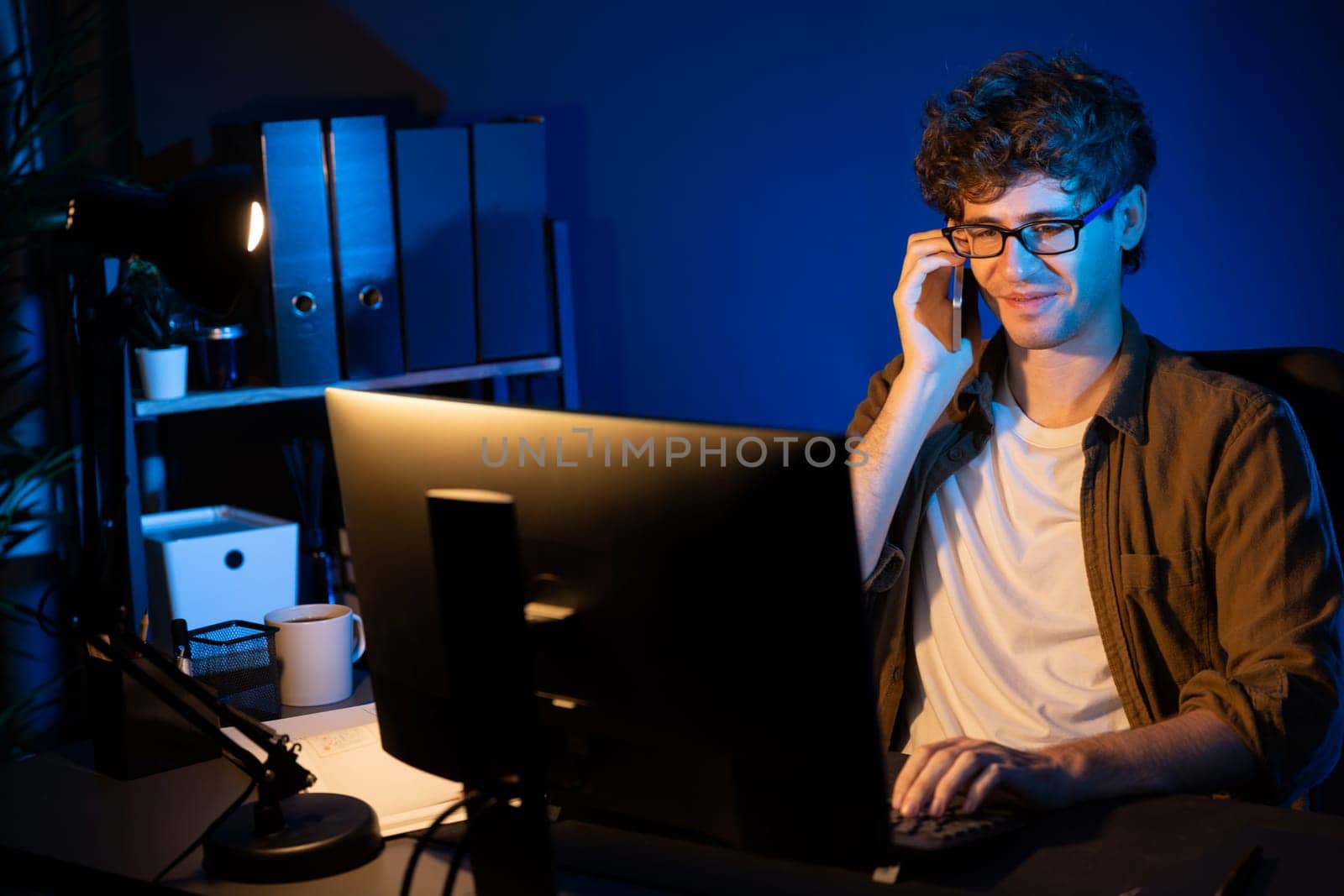 Young businessman calling on smartphone to customer or coworker, looking at computer screen of creative presentation surround work desk at neon modern office. Concept of new next project. Gusher.