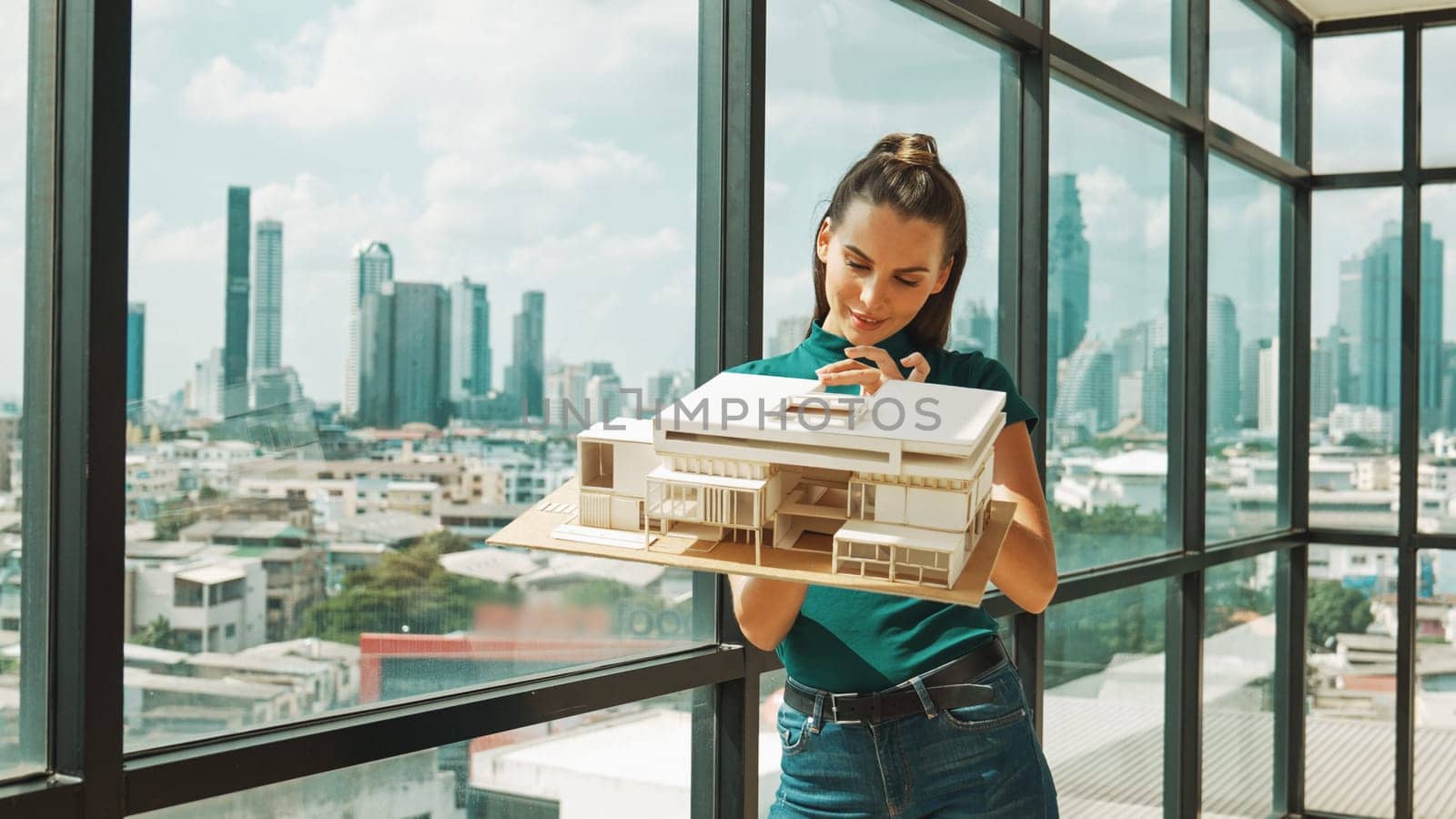 Young smart architect engineer holds architectural model while inspect house model. Professional interior designer checking house construction while standing near window with city view. Tracery