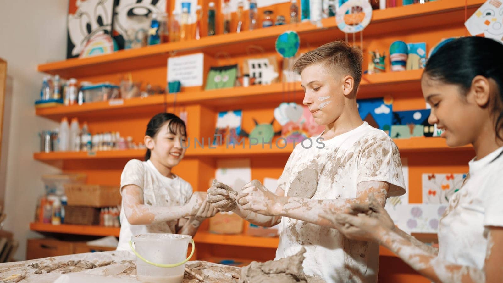 Group of happy diverse children playing clay while wearing muddy shirt at art lesson. Cute highschool girl playing with dough while putting clay on happy boy shirt at pottery workshop. Edification.