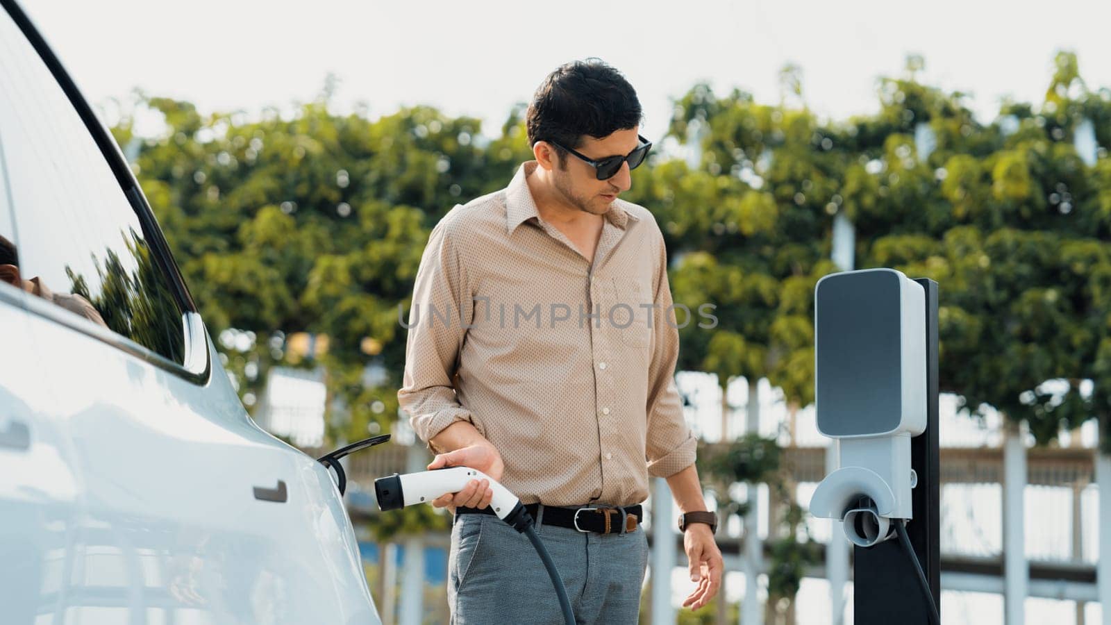 Young man recharge electric car's battery from charging station in outdoor green city park. Rechargeable EV car for sustainable environmental friendly urban travel. Panorama Expedient