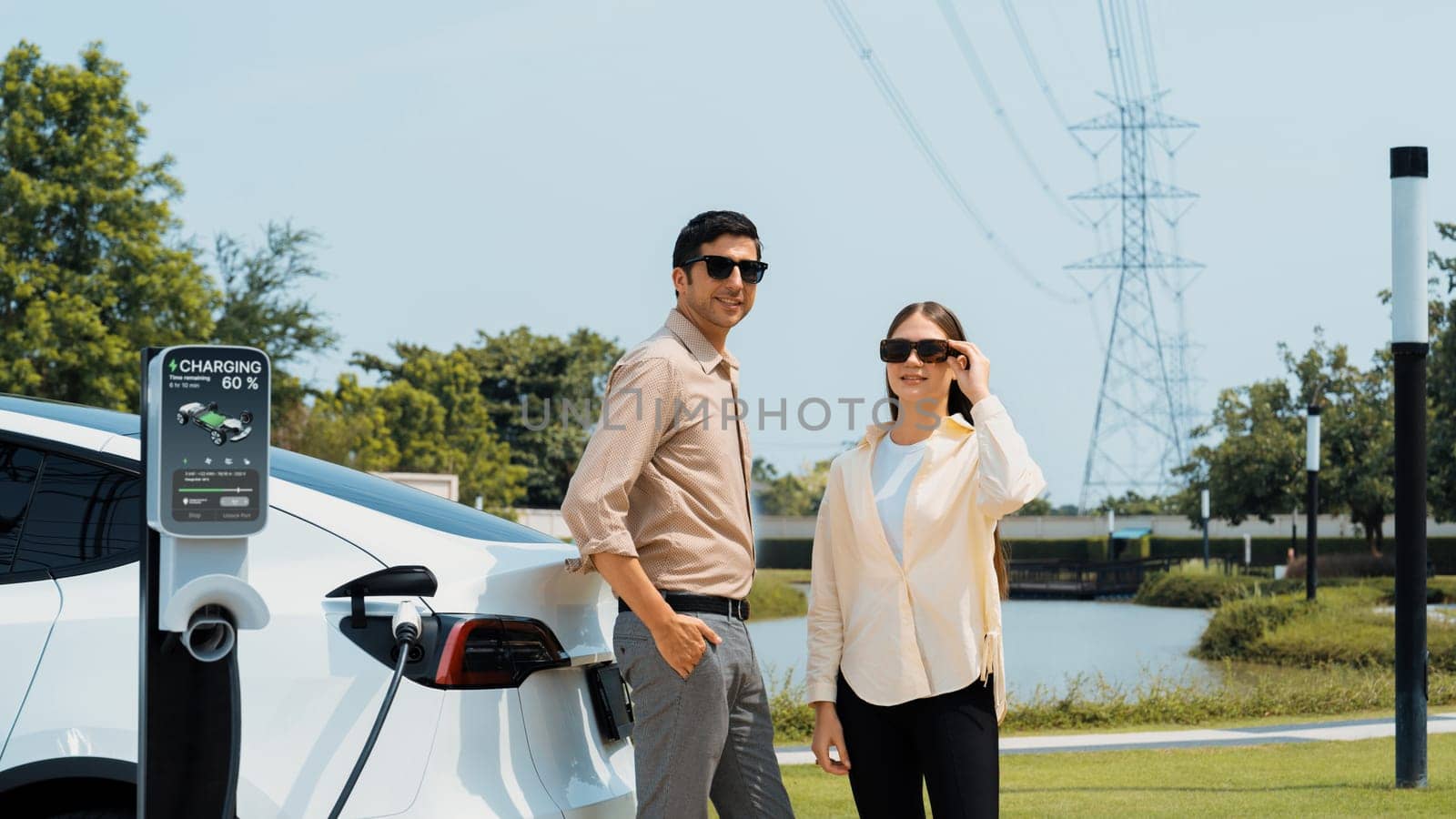 Young couple recharge EV car battery at charging station connected to power grid tower electrical industrial facility as electrical industry for eco friendly vehicle utilization. Expedient