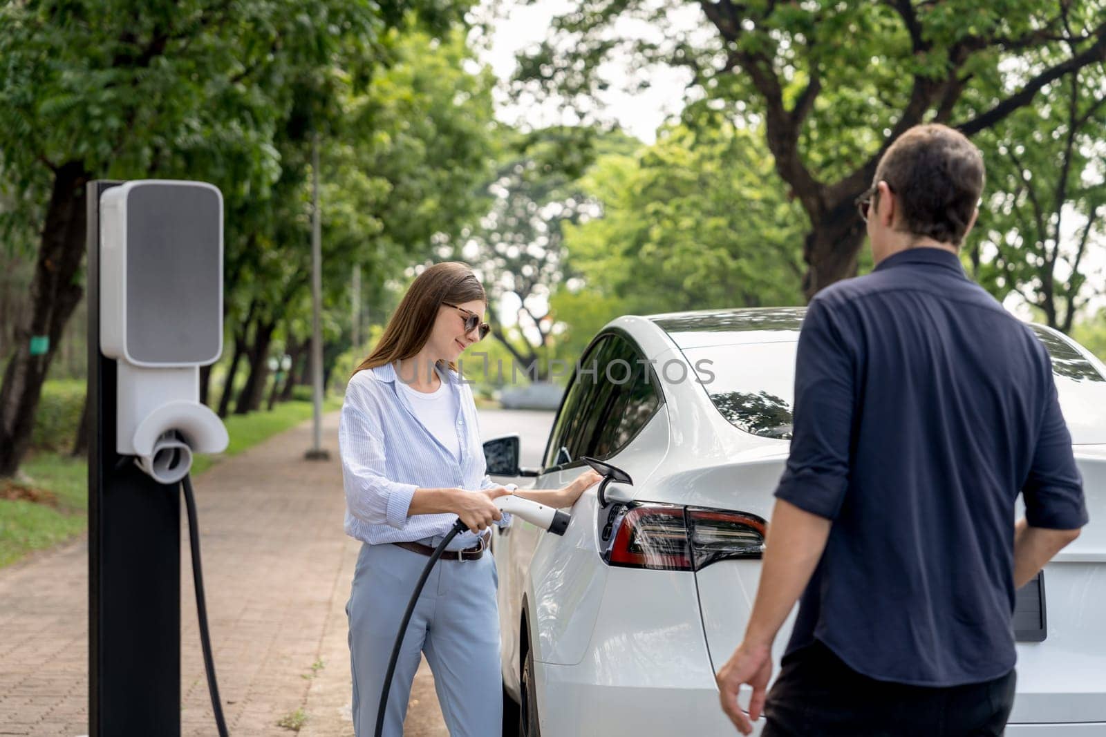 Lovely young couple wearing sun glasses recharging battery for electric car during road trip travel EV car in natural forest or national park. Eco friendly travel during vacation and holiday. Exalt